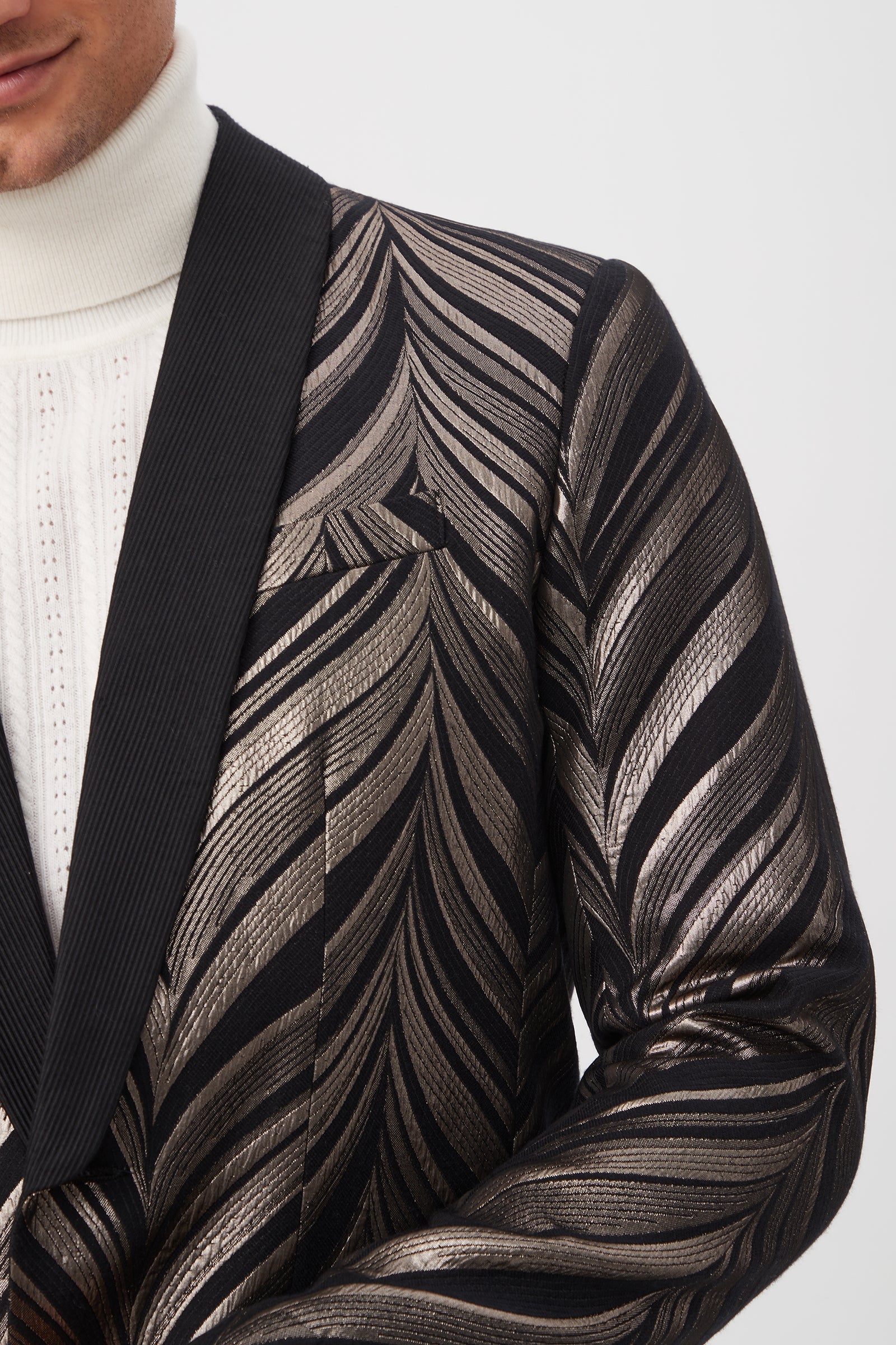 A close-up of a person wearing the Alfred Blazer - Descanso Chevron Jacquard, featuring a black and silver metallic chevron pattern with black lapels, layered over a white ribbed turtleneck. The face is partially out of frame.