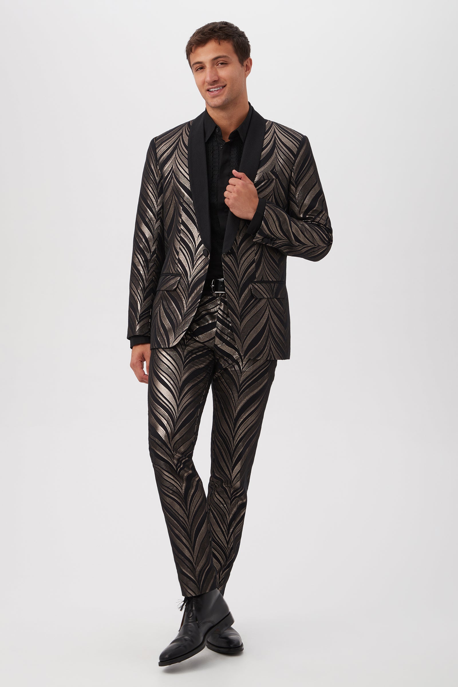 A man smiles in the Alfred Blazer – Descanso Chevron Jacquard, featuring a shiny black finish with bold silver leaf patterns, paired with a black dress shirt and black dress shoes against a white background.