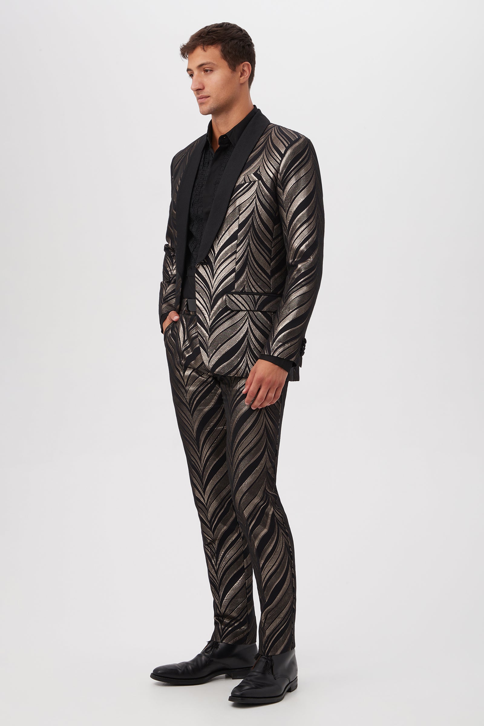 A man stands against a plain white background wearing the Alfred Blazer in Descanso Chevron Jacquard, paired with a black shirt and dress shoes. The metallic, leaf-like pattern of the blazer stands out as he looks to the side, hands in pockets.