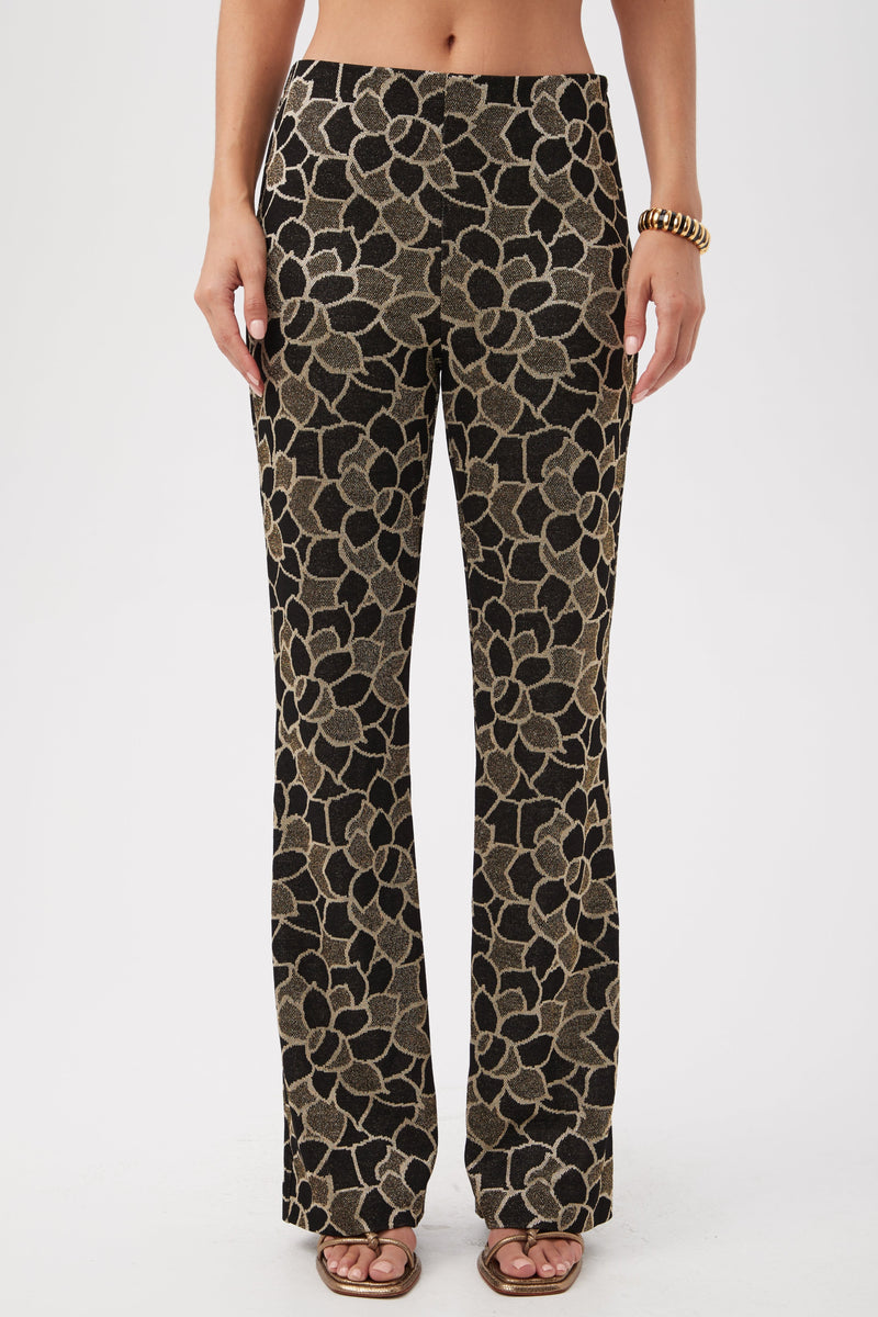 A person wearing Jacoba Metallic Knit Jacquard Bootcut Pants in multi, featuring a bold floral pattern, stands in sandals and a gold bracelet against a plain white background.