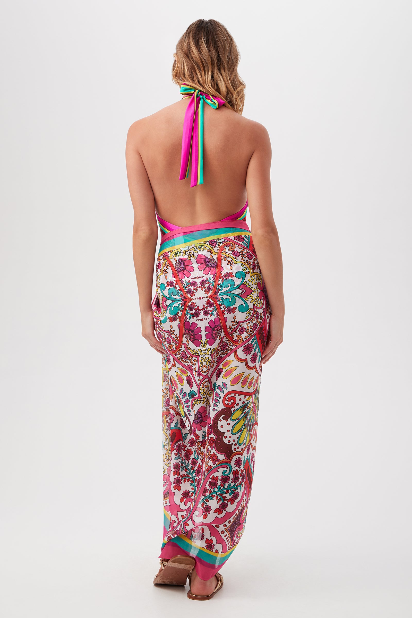 A woman faces away, modeling the Hawaiian Garden Pareo Swim Cover-Up—a resort-ready, halter-neck, backless dress with a colorful floral and paisley print. It features pink, green, and yellow straps tied at the neck and waist. She wears sandals.