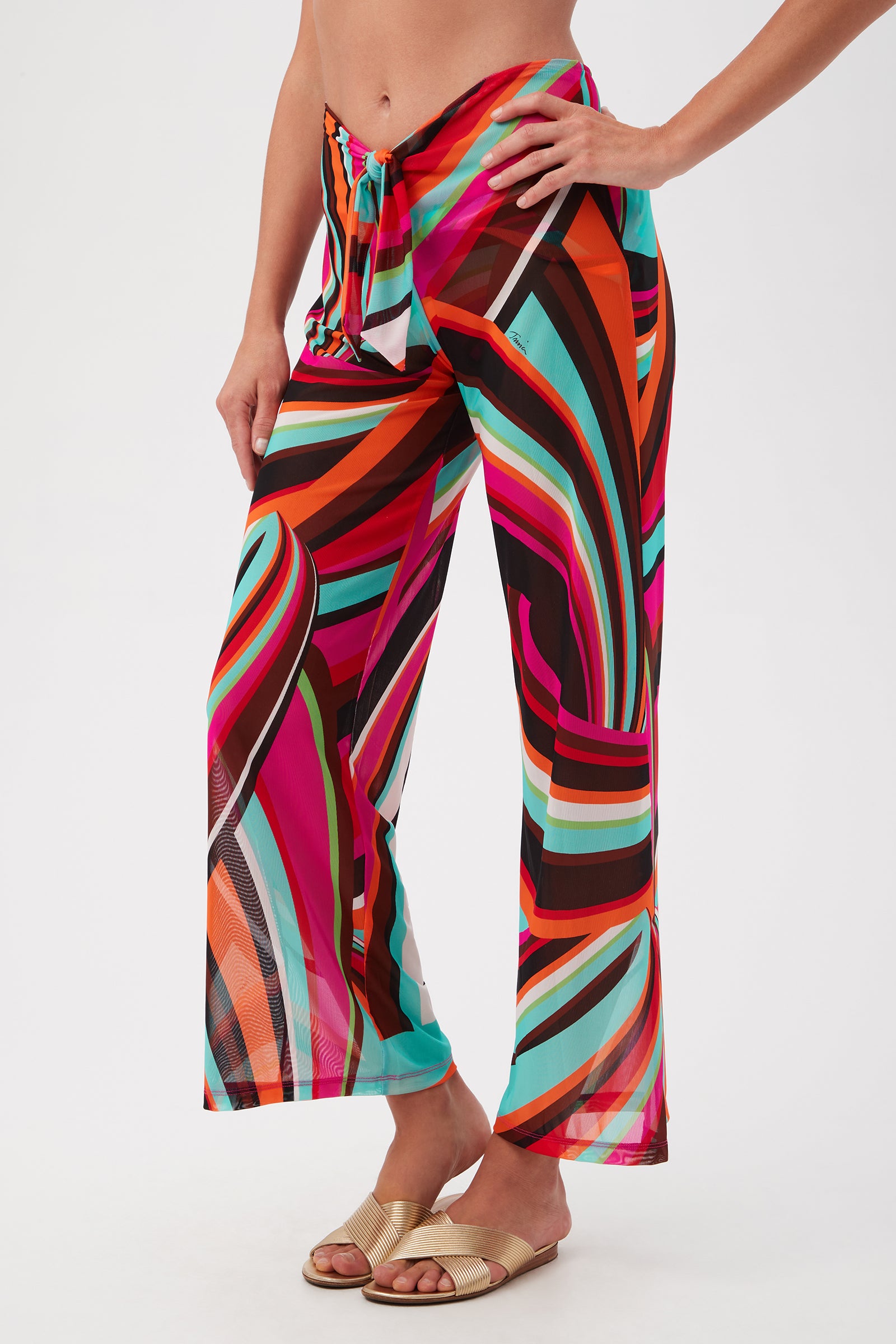 A person wearing Swoop Mesh Tie-Front Wide-Leg Cover-Up Pants with a vibrant abstract tropical pattern in pink, orange, black, teal, and white, paired with gold slide sandals and standing against a plain white background.