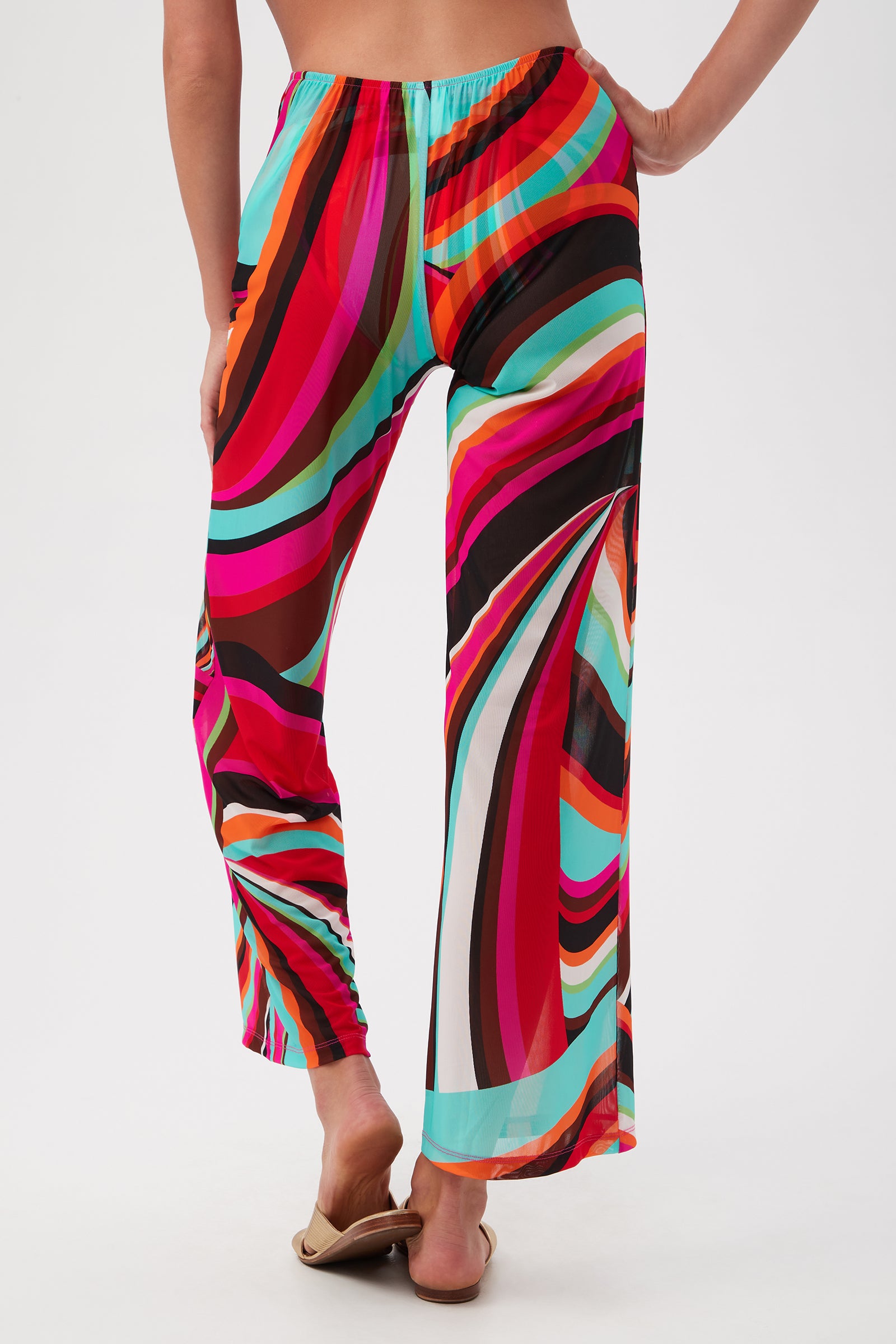 A person stands barefoot on a white background, shown from the back, wearing Swoop Mesh Tie-Front Wide-Leg Cover-Up Pants featuring a vibrant swirl pattern in red, pink, teal, orange, white, and black.