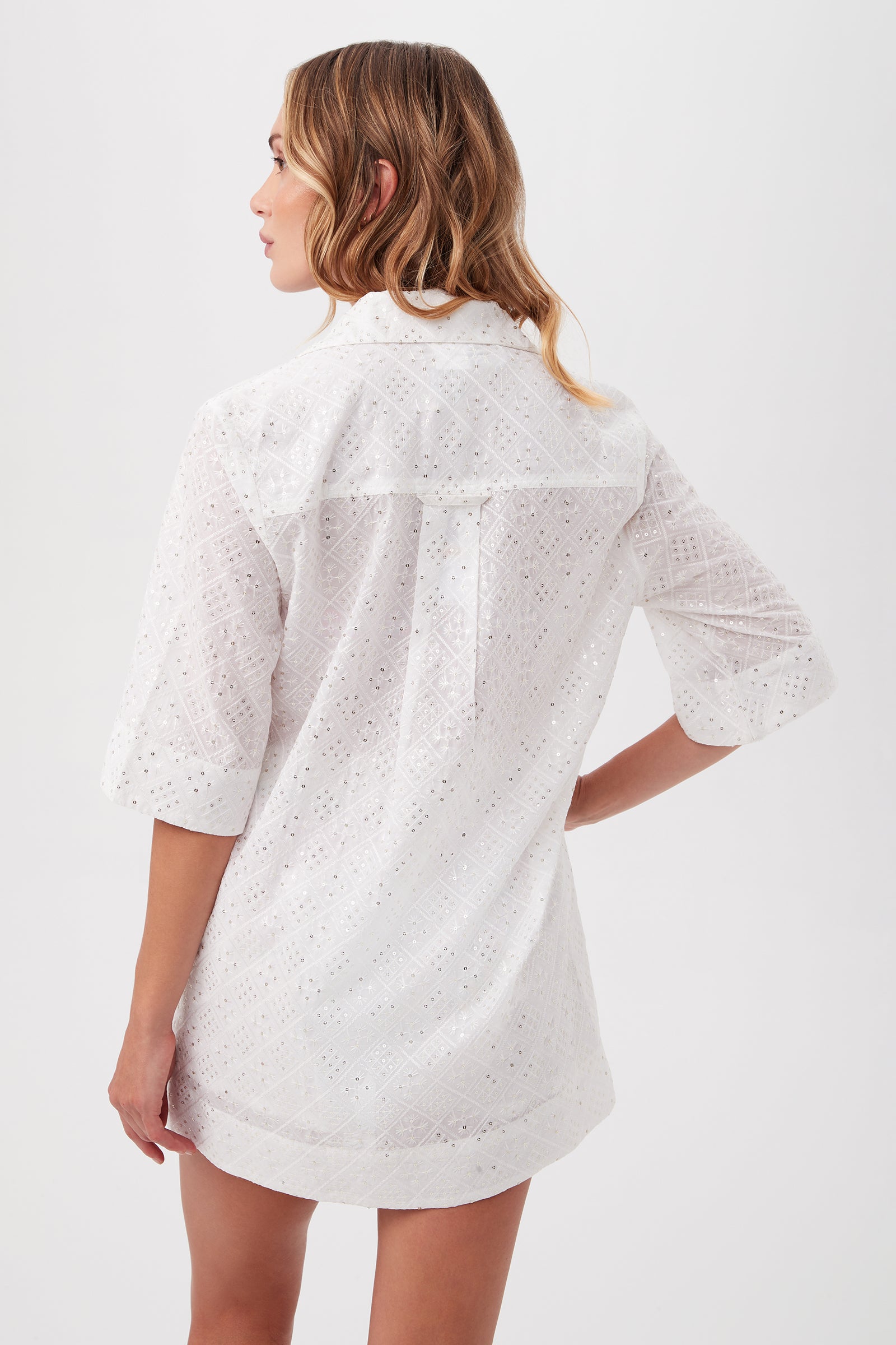 A woman with wavy, light brown hair models the Mazarine Sequin Tunic Beach Cover-Up Shirt With Pockets. She stands facing away from the camera against a plain white background, wearing the white, three-quarter sleeve cover-up.