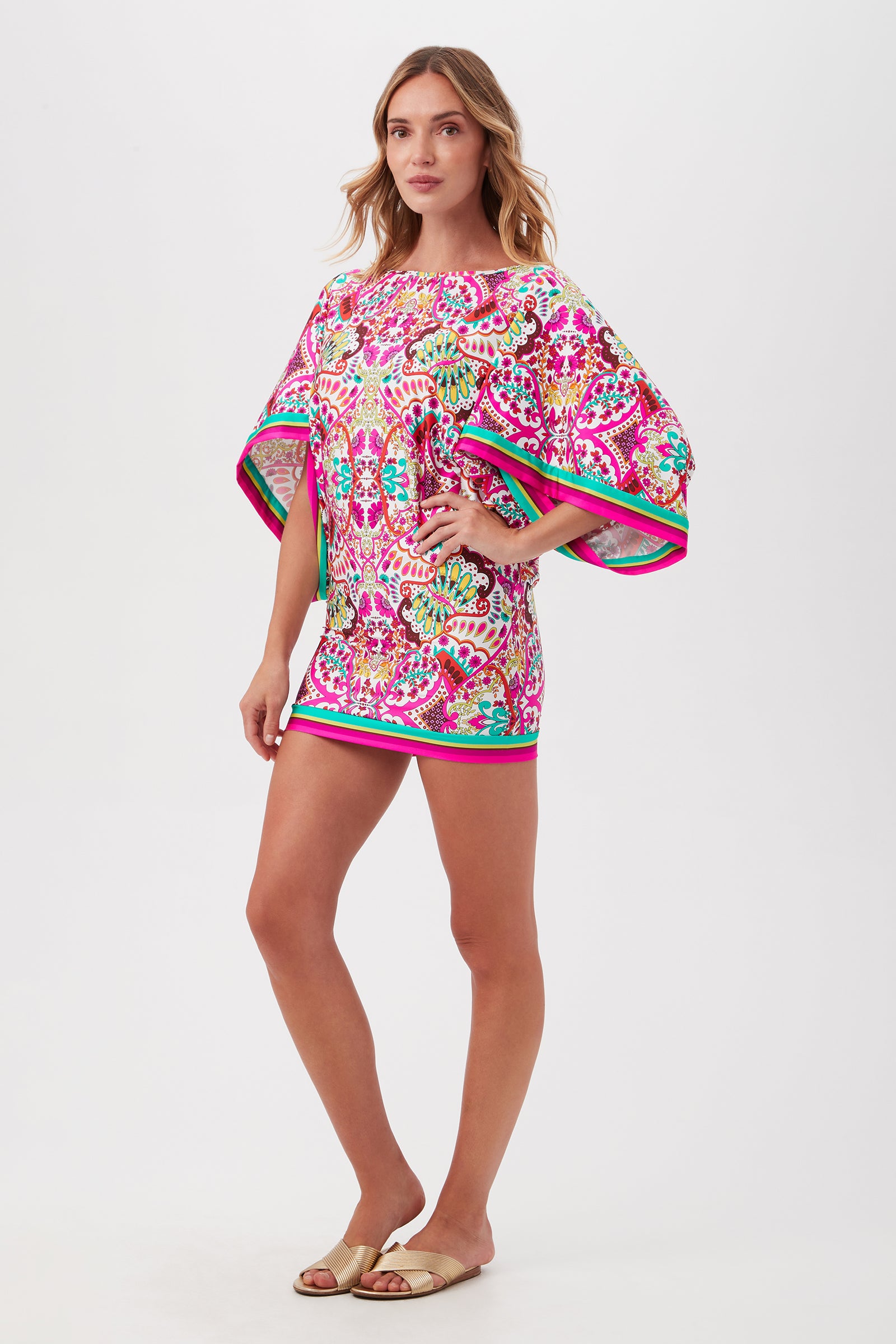 A woman with long wavy hair and beige sandals wears the Hawaiian Garden Boatneck Tunic Swim Cover-Up Dress in vibrant pink, green, and white patterns, while a plain white background highlights her stylish look.