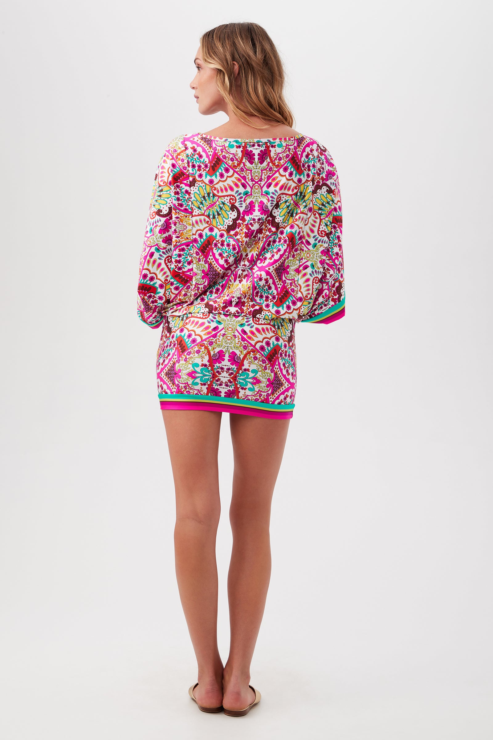 A woman stands barefoot against a white background, wearing the Hawaiian Garden Boatneck Tunic Swim Cover-Up Dress. The long sleeves and pink, green, and yellow patterns complement her wavy, shoulder-length hair.