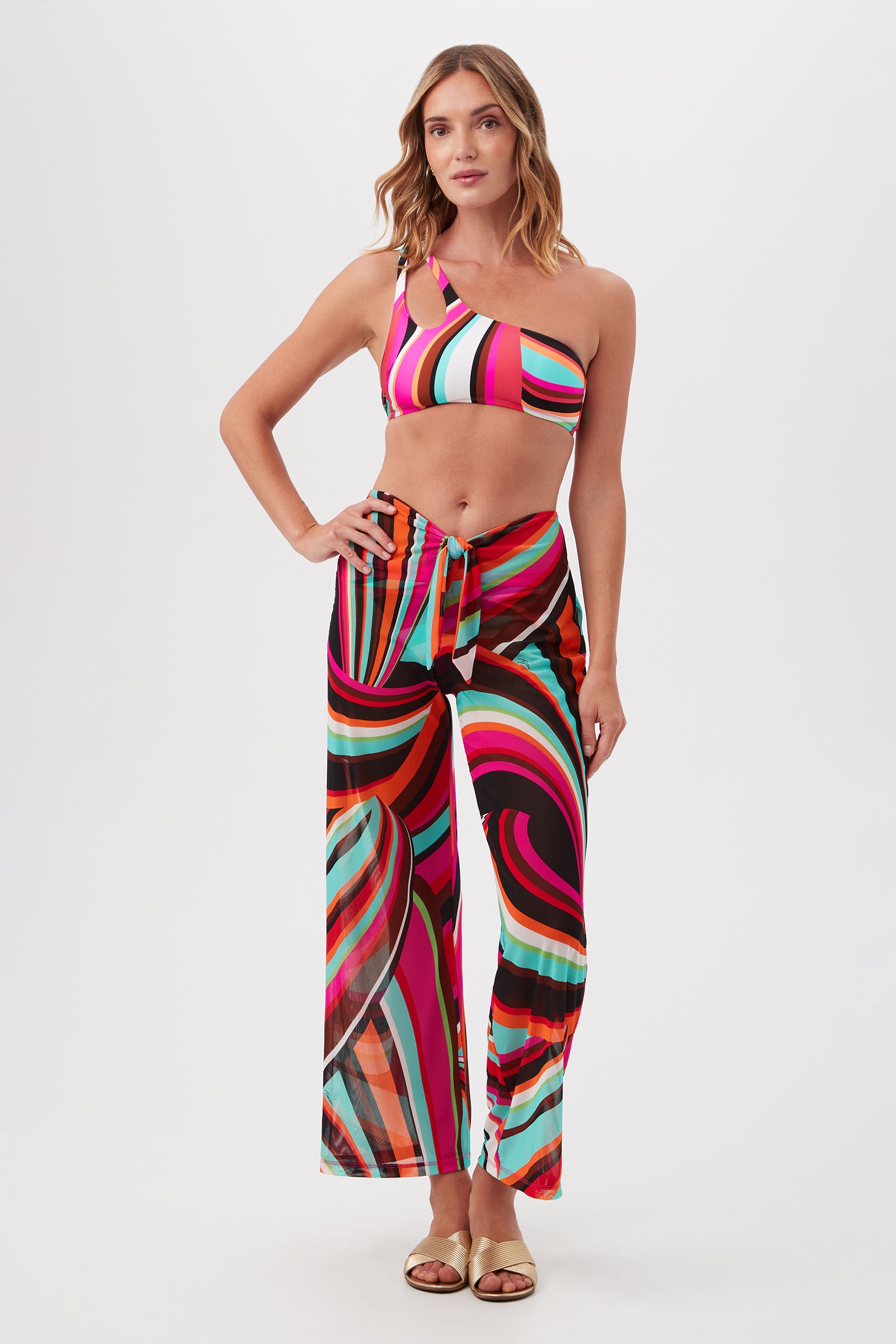 A woman poses confidently in Swoop Mesh Tie-Front Wide-Leg Cover-Up Pants with a colorful tropical print, paired with a matching one-shoulder crop top against a white background. She has light brown hair and wears sandals.