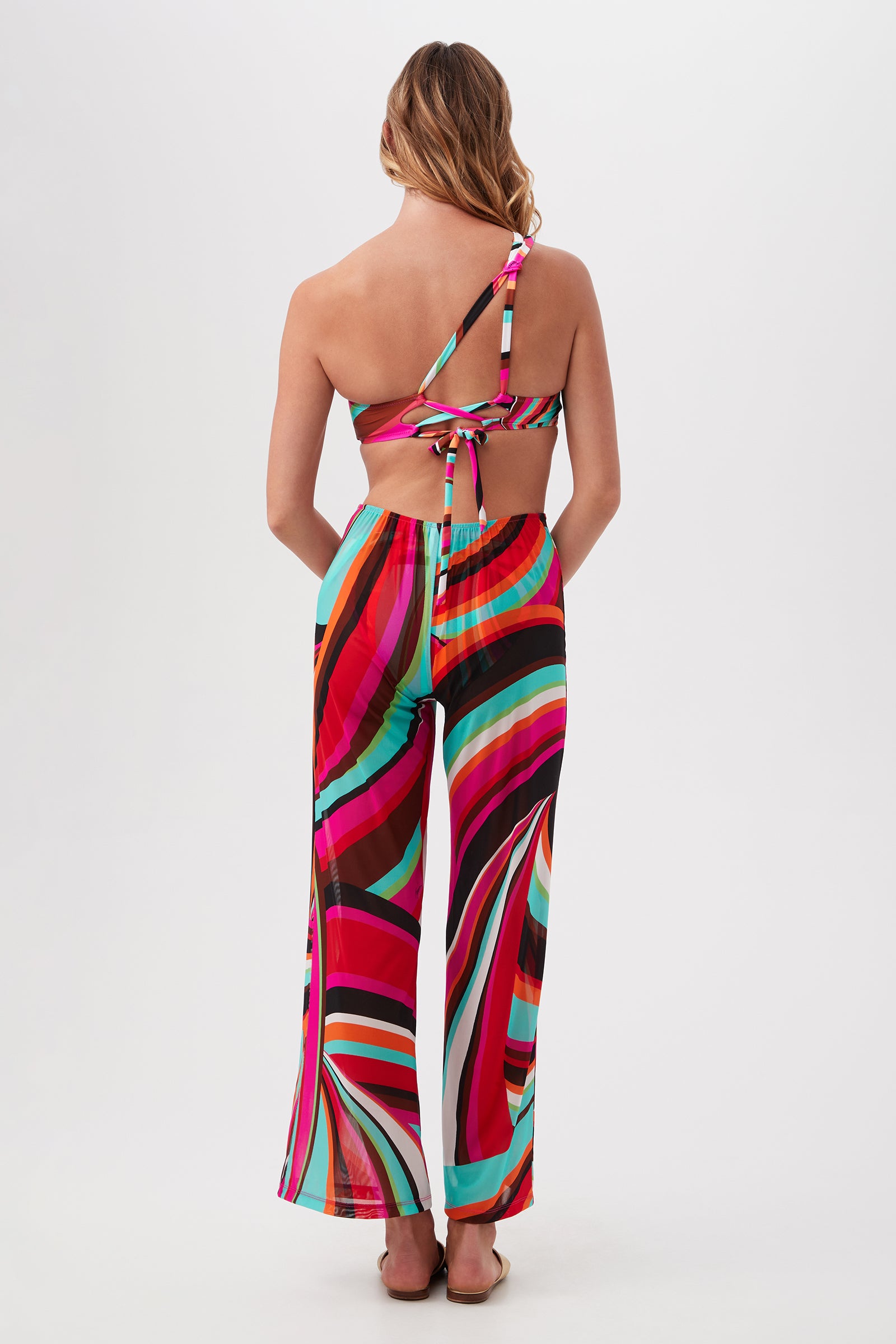 Woman stands with her back to the camera, wearing colorful Trina Turk swimwear and Swoop Mesh Tie-Front Wide-Leg Cover-Up Pants in vibrant red, pink, turquoise, black, and white.