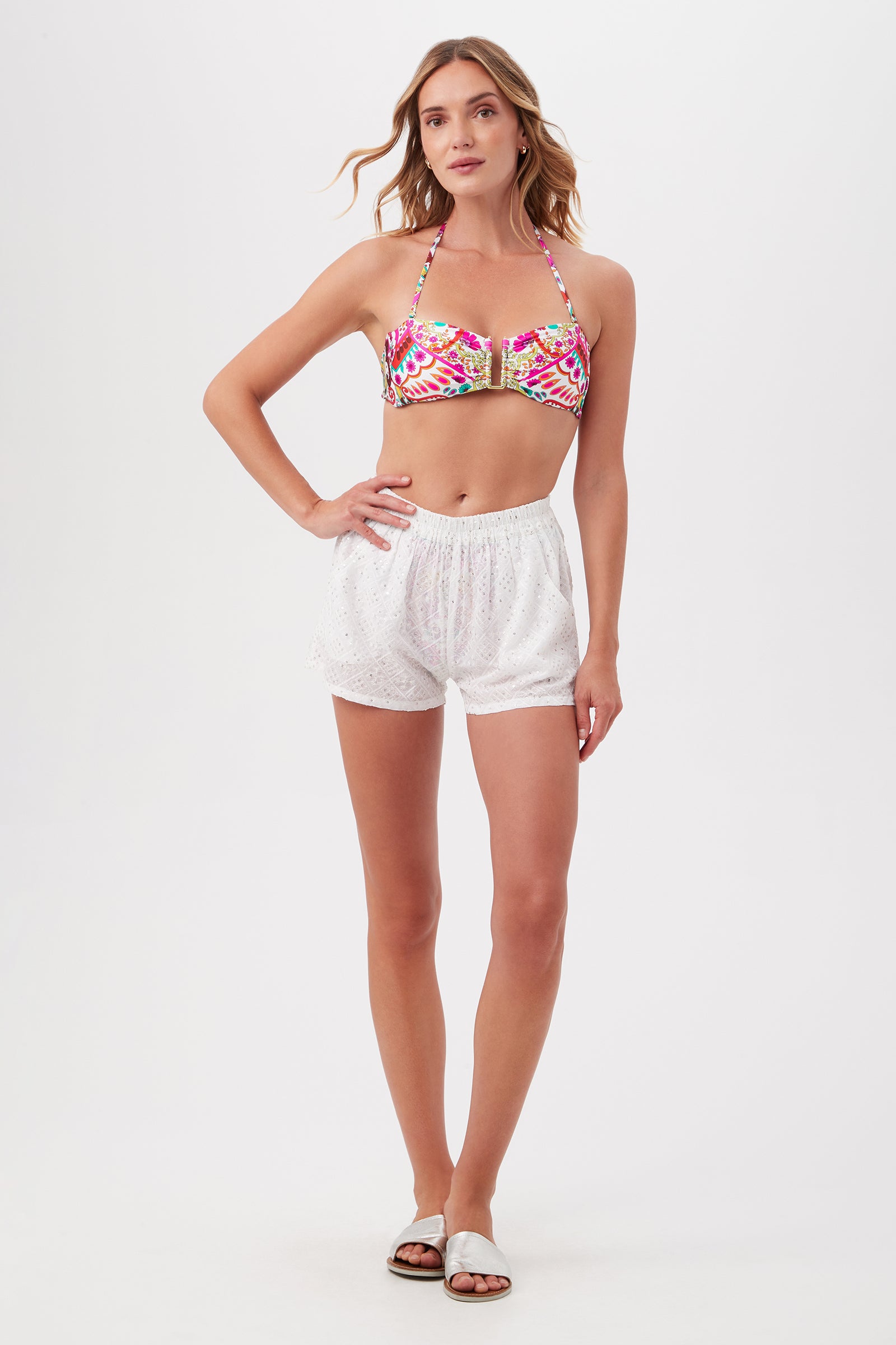 A woman stands against a white background wearing a colorful bikini top, Mazarine Sequin Cover-Up Shorts With Pockets in white, and white sandals, her hair loose and one hand on her hip—perfect for a stylish beach day.