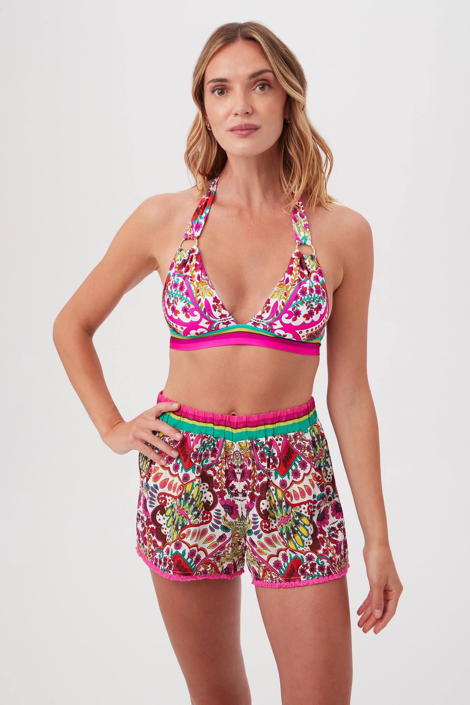 A woman with shoulder-length blonde hair and a neutral expression stands against a white background wearing the Trina Turk Hawaiian Garden Ring Halter Swim Top in Multi color with matching high-waisted shorts.