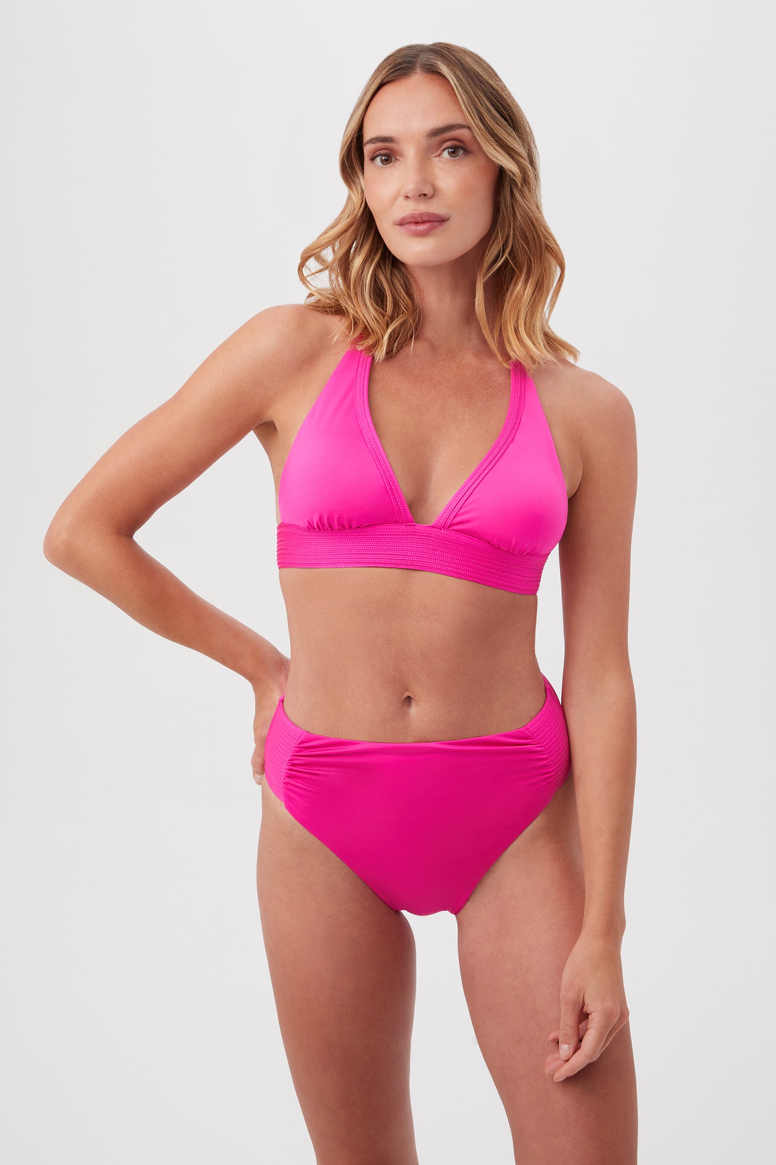 A woman with shoulder-length wavy hair and a confident expression models the Monaco Pleated Halter Triangle Top in bright pink bikini style, paired with high-waisted bottoms against a plain white background.