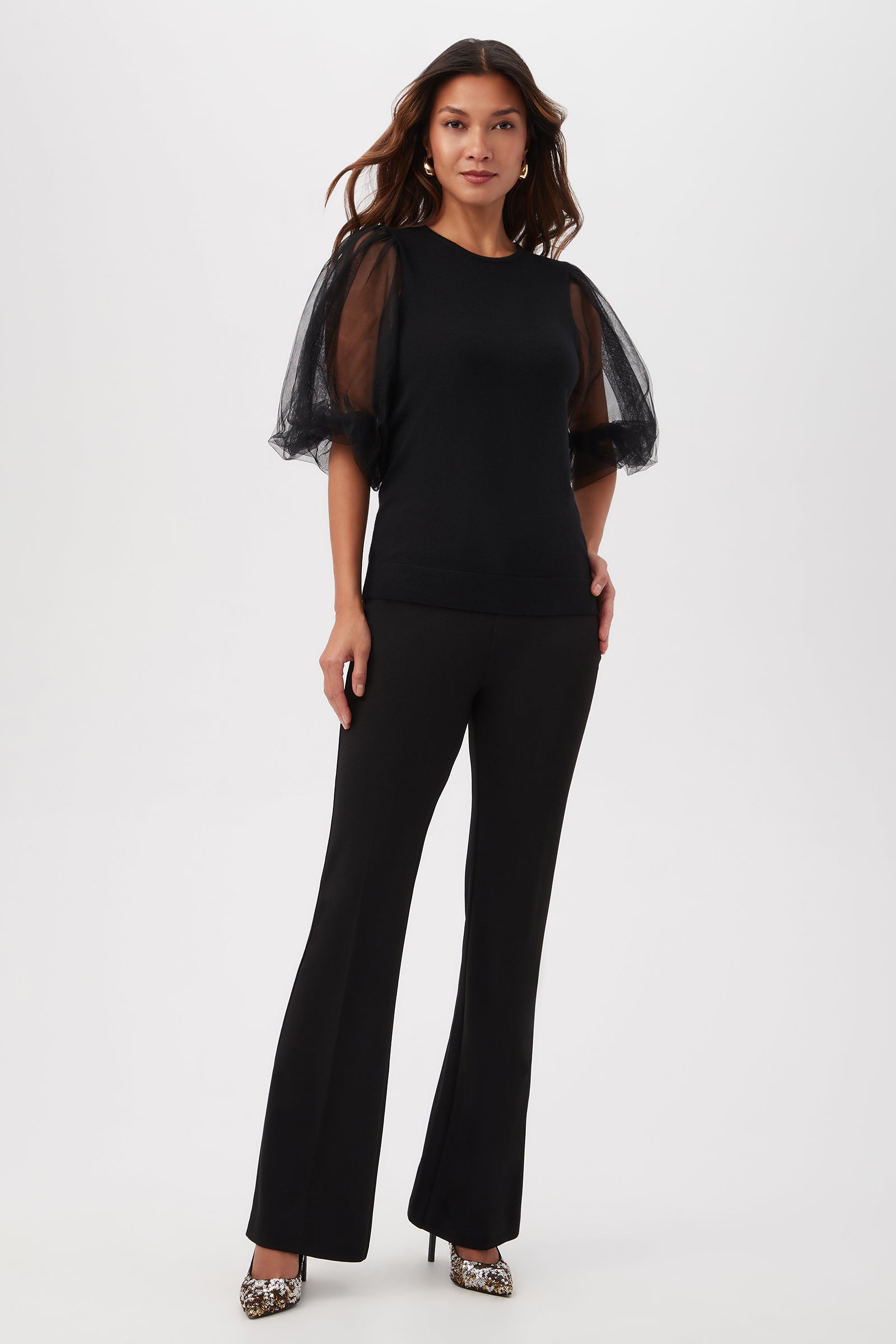A woman with long, wavy hair and a neutral expression stands against a white background wearing the Rhea 3 Merino Wool Crewneck Sweater with sheer short sleeves, paired with black flared pants and leopard-print high heels.