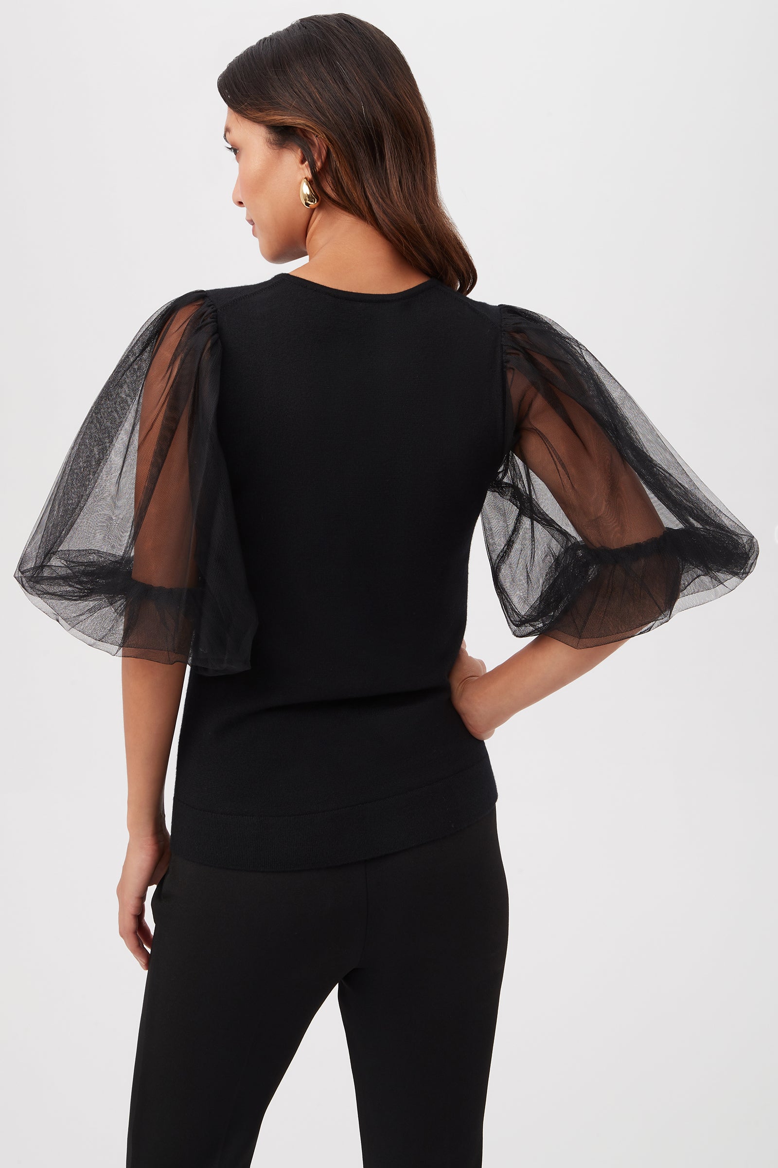 A woman with long brown hair is seen from behind wearing the Rhea 3 Merino Wool Crewneck Sweater with sheer short sleeves and black pants, against a plain light background.