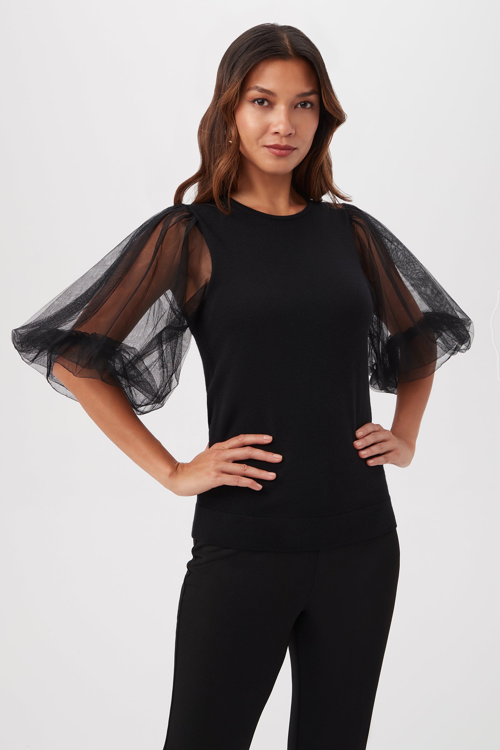 A woman poses confidently in the Rhea 3 Merino Wool Crewneck Sweater with sheer short sleeves in black and matching pants, set against a plain light background.