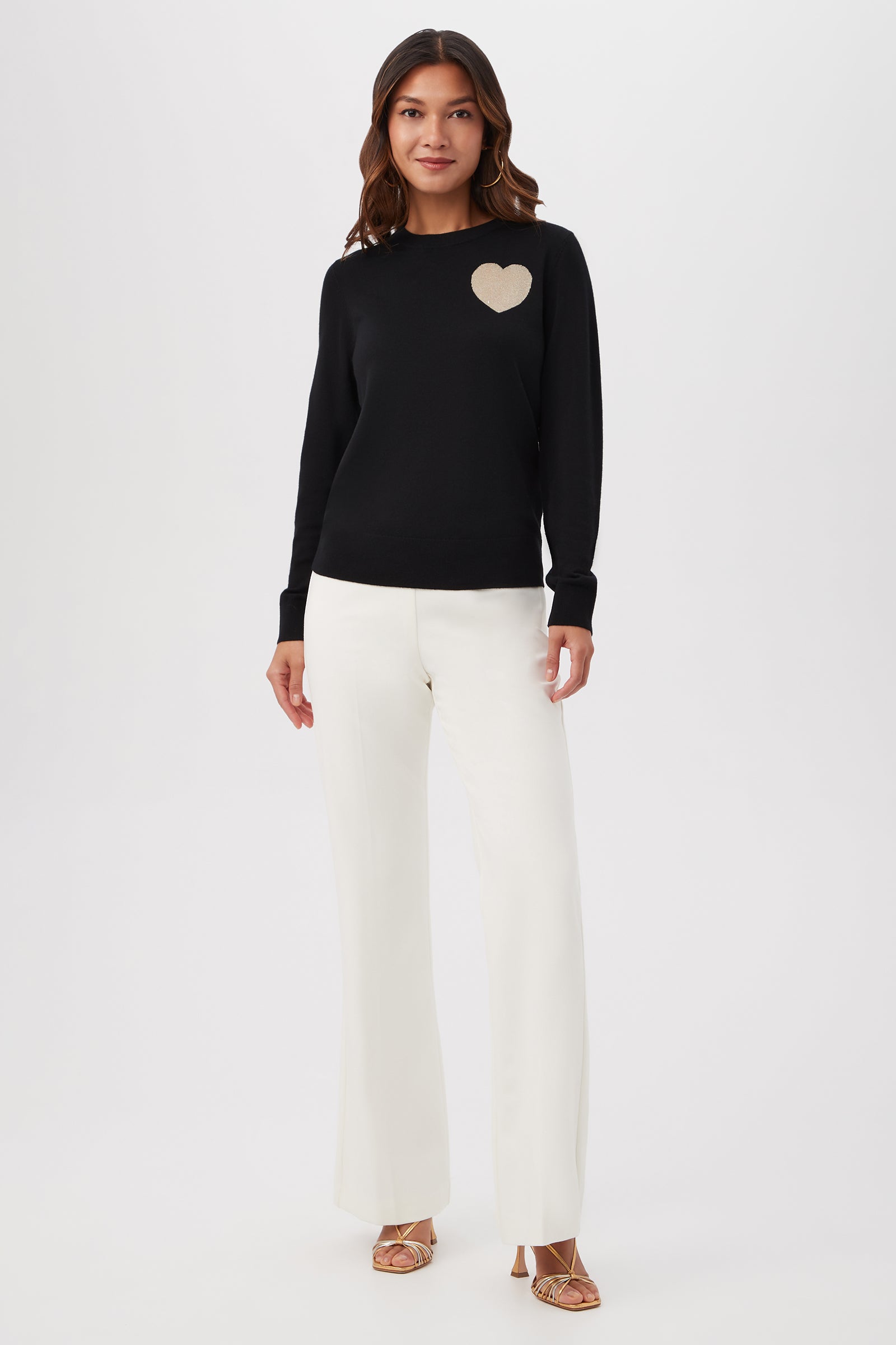 A woman with wavy brown hair and a slight smile stands against a white background wearing the Gold Heart Merino Wool Crewneck Sweater, featuring a beige heart on the chest, along with white wide-leg pants and beige open-toe heels.