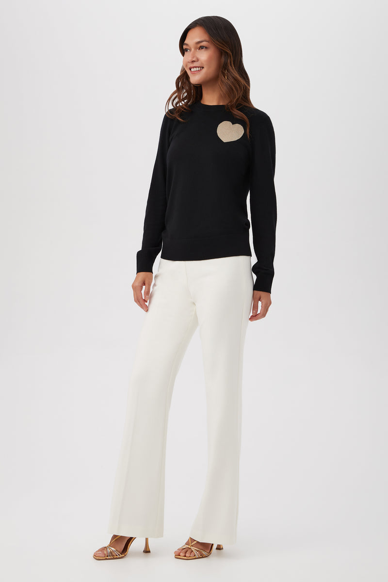 A woman smiles, wearing the Gold Heart Merino Wool Crewneck Sweater, white wide-leg pants, and gold open-toe heels against a plain white background.