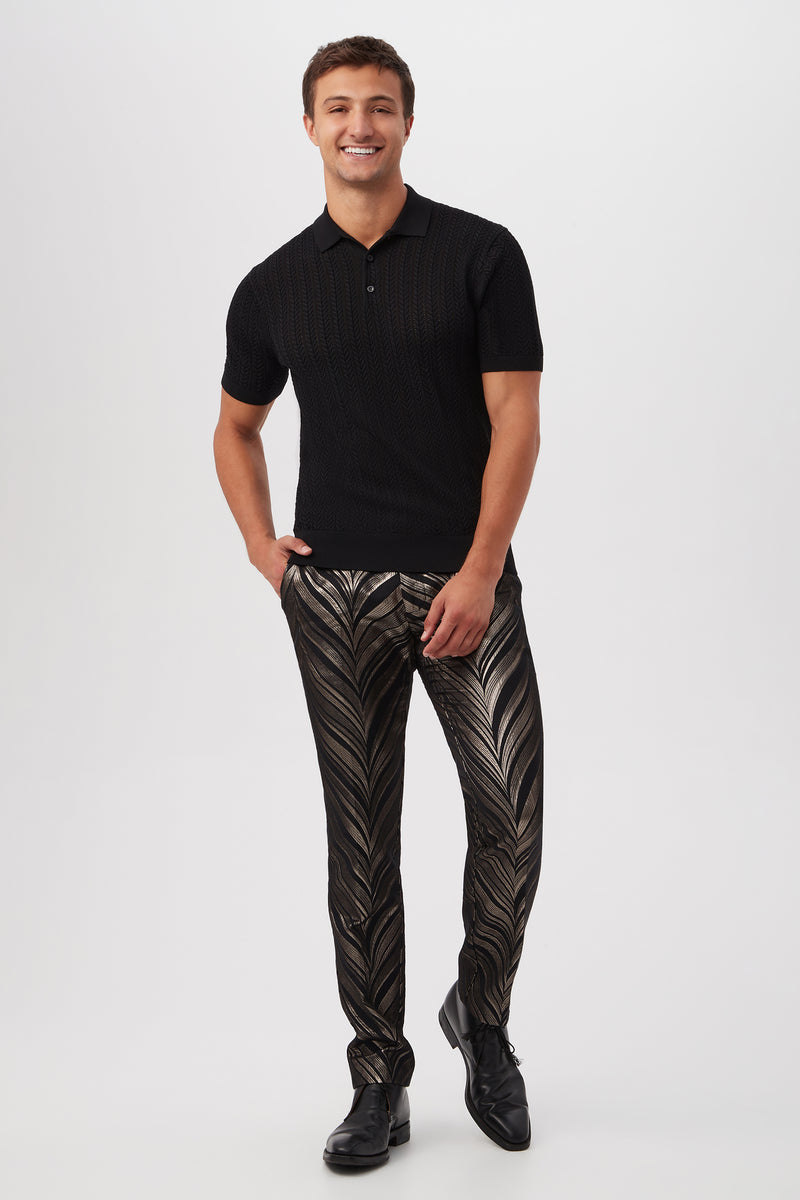 A man smiles against a plain white background, wearing the Maxton Short-Sleeve Polo - Cotton Modal in black, paired with shiny, patterned black and gold dress pants and black dress shoes.