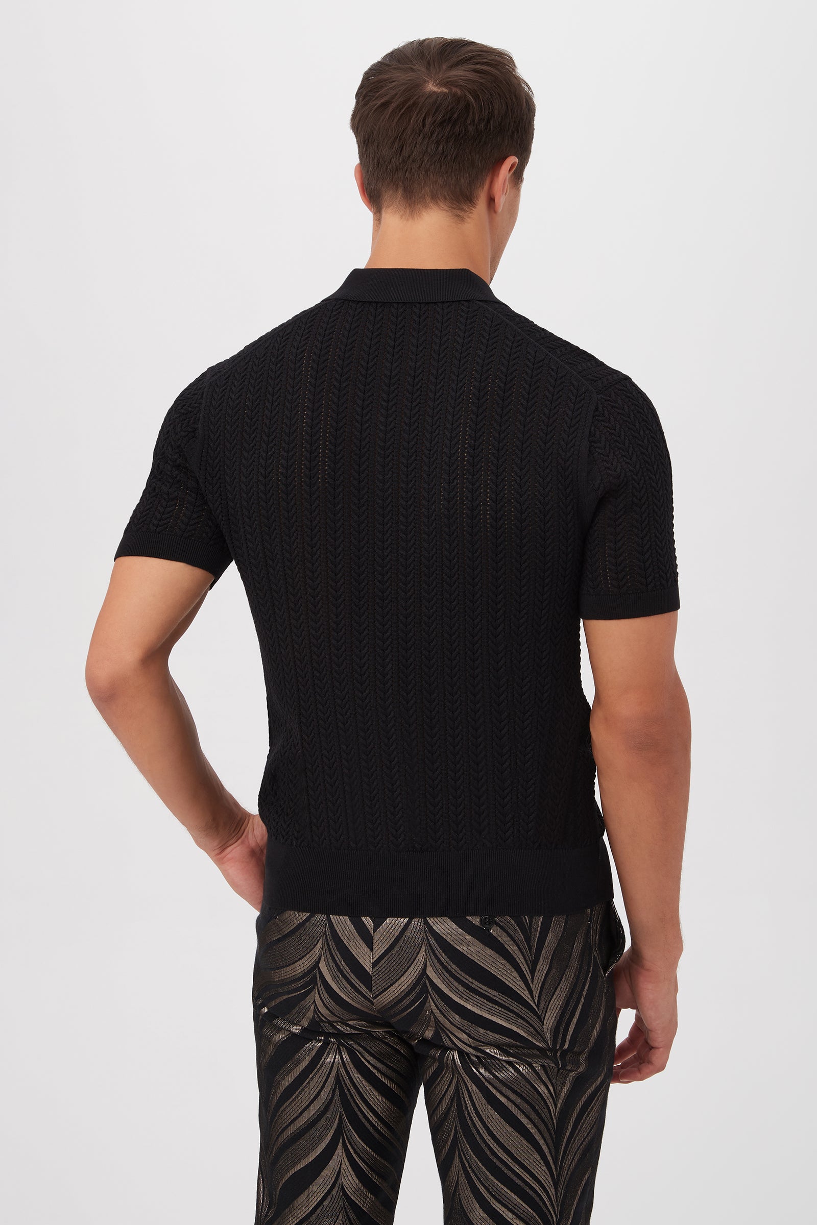 A man stands with his back turned, wearing the Maxton Short-Sleeve Polo - Cotton Modal in black and metallic black pants with a shiny leaf pattern, set against a plain white background.