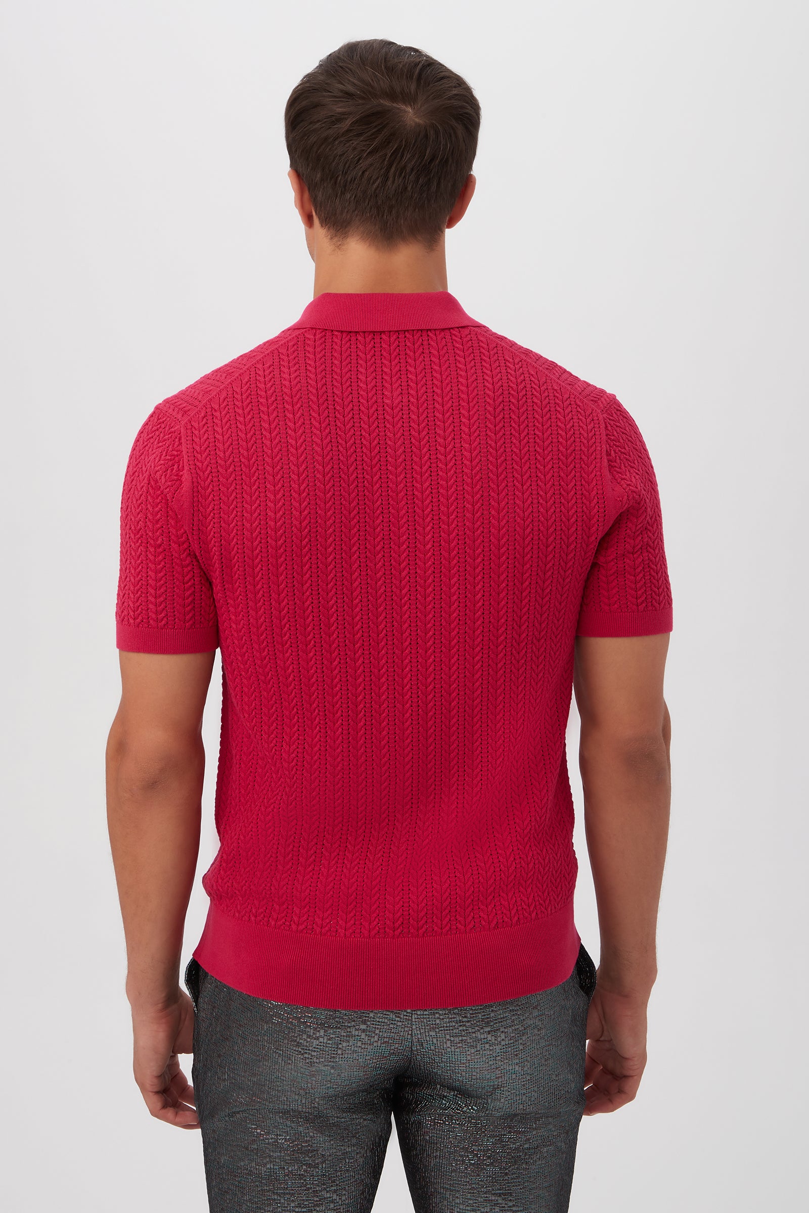 A man with short brown hair stands with his back to the camera, wearing the Maxton Short-Sleeve Polo - Cotton Modal in bright red and dark, shiny pants against a plain light background.