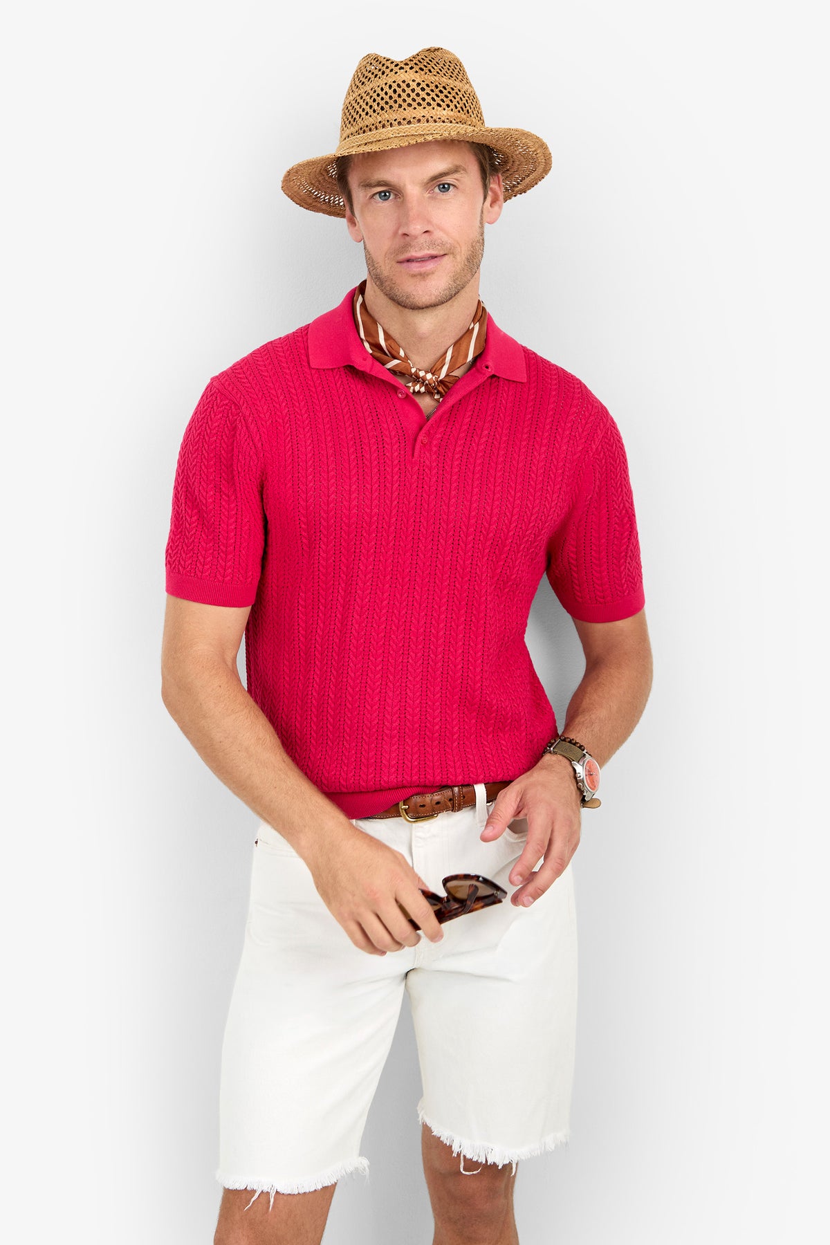 Man wearing a straw hat, Maxton Short-Sleeve Polo - Cotton Modal in red, white frayed shorts, brown belt, patterned neck scarf, and holding sunglasses against a plain light background.