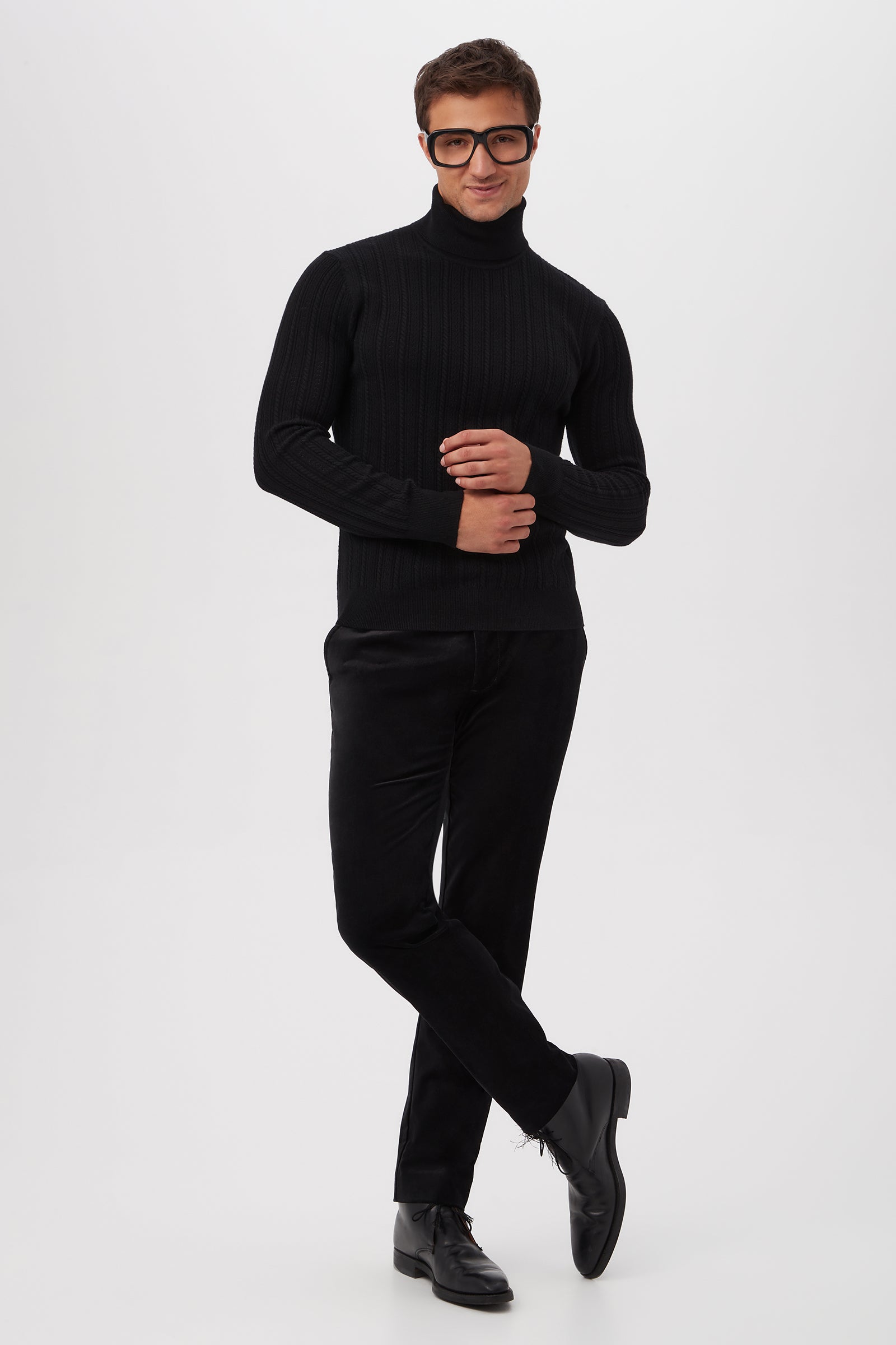 A man poses confidently in the Oberon Turtleneck Sweater - Merino Wool, styled with black pants and shoes. He wears large, dark-rimmed glasses and smiles slightly against a plain white background, standing with one leg crossed and hands together.