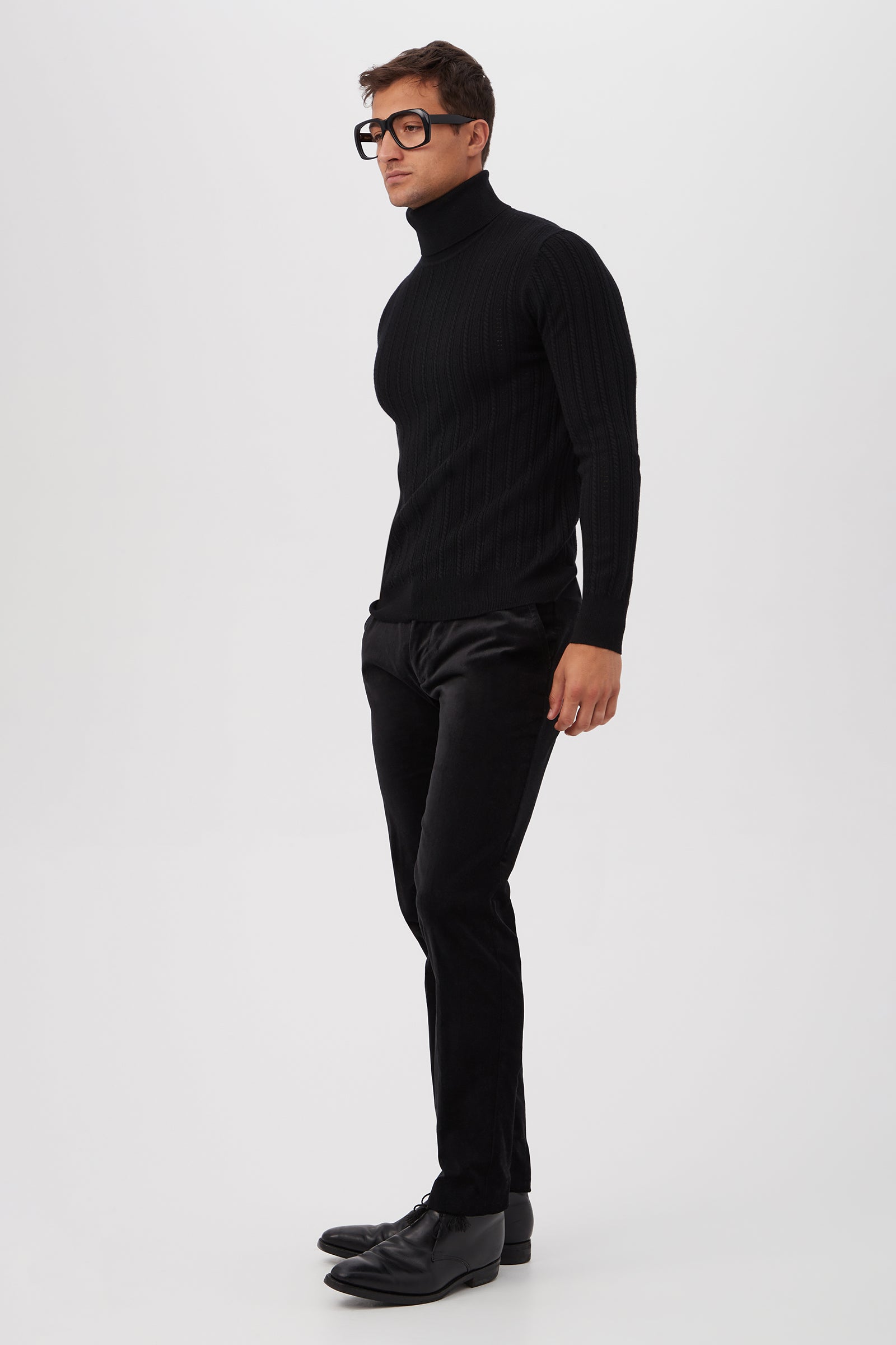 A man wearing the Oberon Turtleneck Sweater - Merino Wool, black slim-fit pants, and black shoes stands against a plain white background. He has short hair and wears large, black-rimmed glasses.