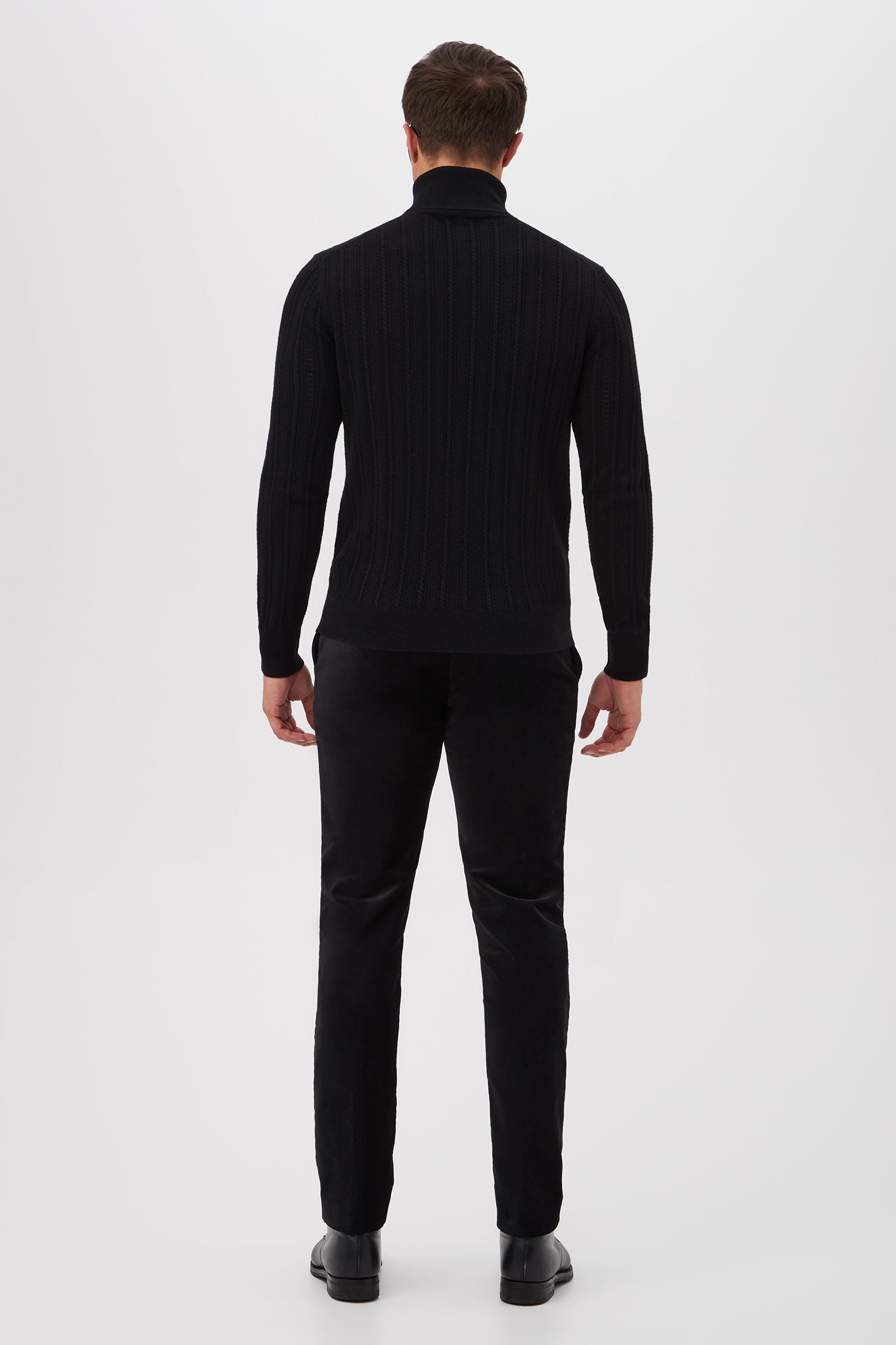 A man faces away from the camera against a white background, wearing the Oberon Turtleneck Sweater - Merino Wool in black, paired with black pants and shoes.