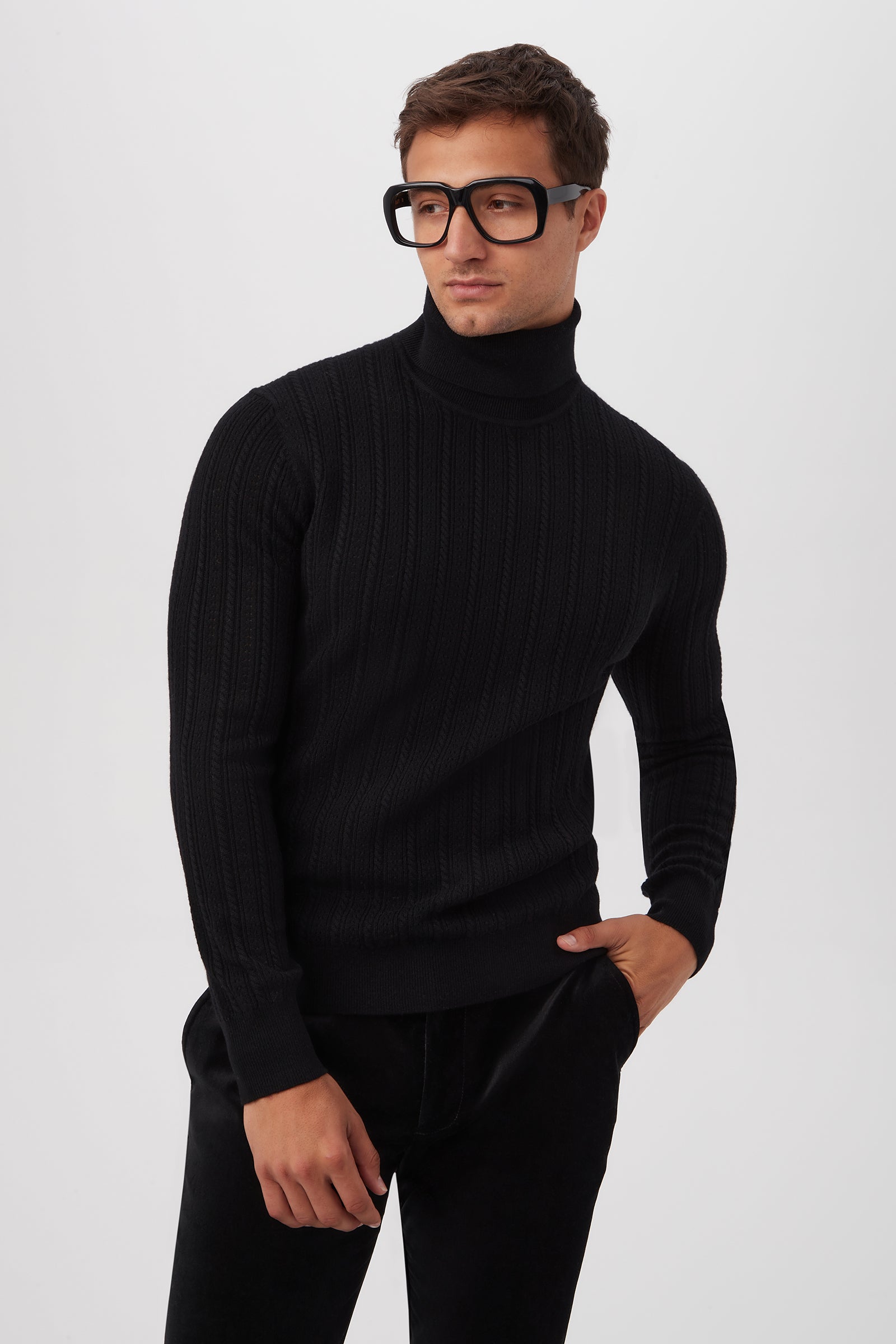 A man stands against a plain white background, wearing the black Oberon Turtleneck Sweater made from merino wool and large black glasses, with one hand in his pocket and looking slightly to the side.