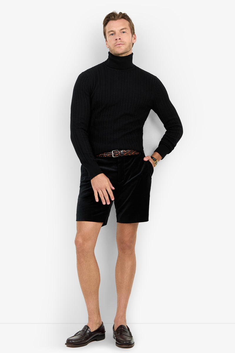 A man poses against a white background wearing the Oberon Turtleneck Sweater - Merino Wool, black shorts, a braided belt, brown loafers, and a wristwatch with one hand resting on his leg.