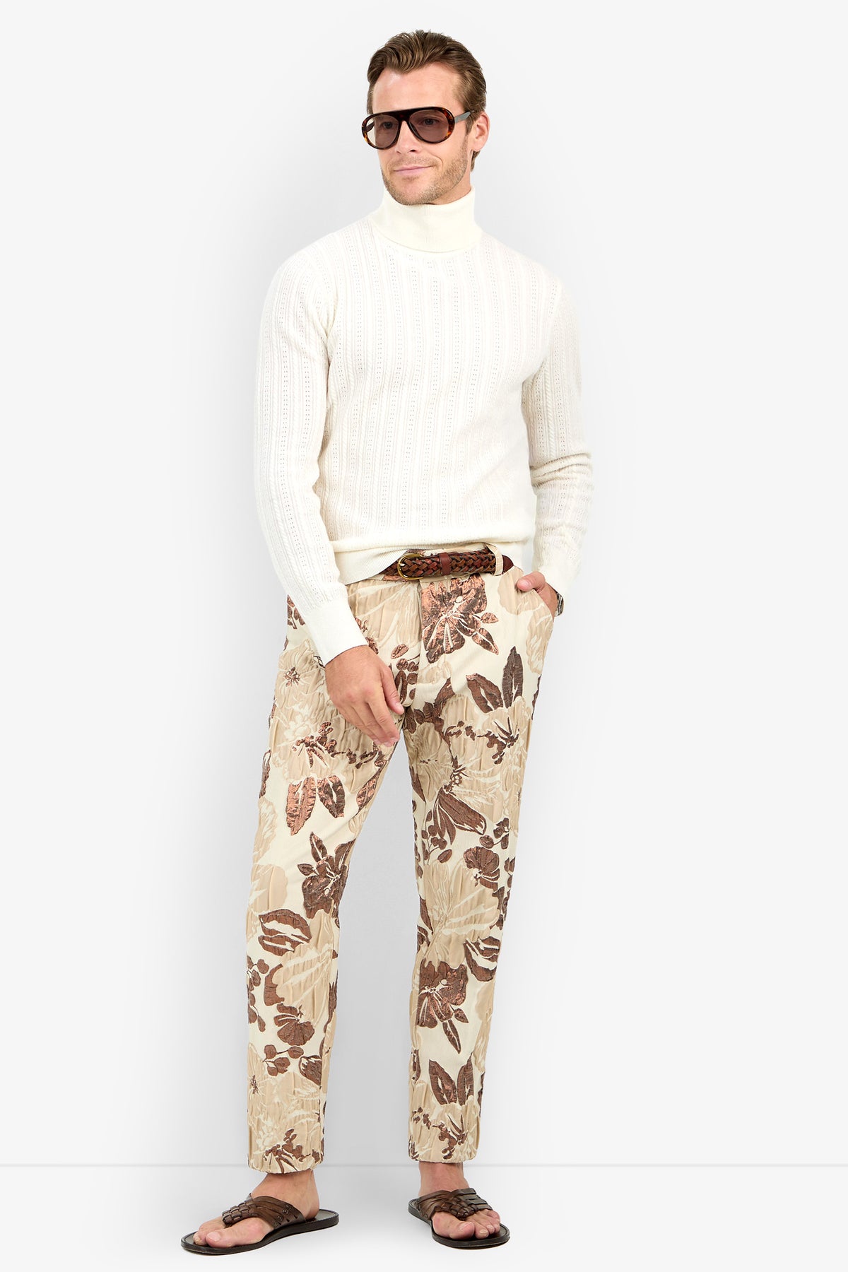 A man in an Oberon Turtleneck Sweater - Merino Wool, sunglasses, beige floral print pants, a brown belt, and flip-flops stands with one hand in his pocket against a plain background.