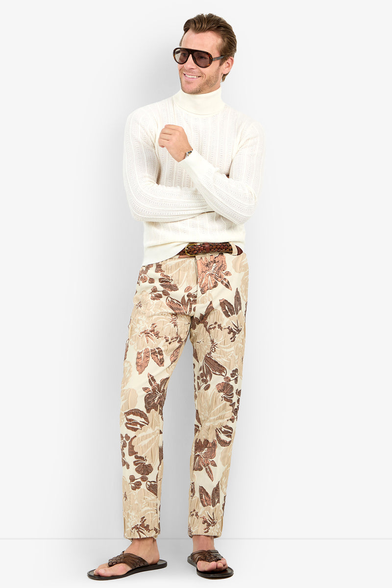 A man wearing sunglasses, the Oberon Turtleneck Sweater - Merino Wool, beige floral print pants, and brown sandals stands smiling with arms loosely crossed.
