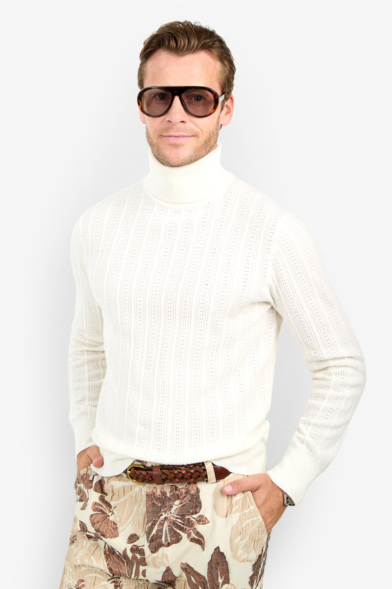 A man wearing the Oberon Turtleneck Sweater - Merino Wool, large brown sunglasses, and patterned beige pants stands with hands in pockets against a plain light background.
