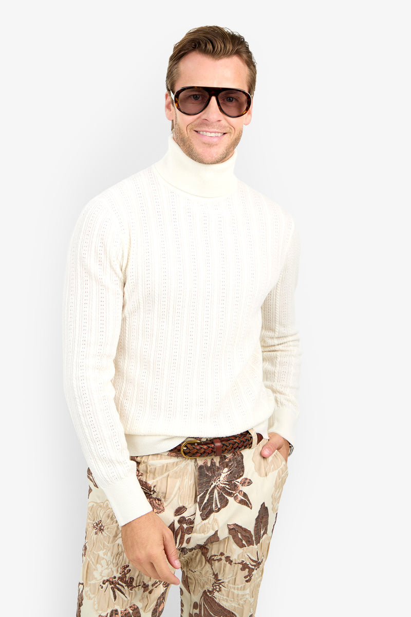 Man wearing Oberon Turtleneck Sweater - Merino Wool, large sunglasses, and beige floral pants, stands smiling against a plain white background.