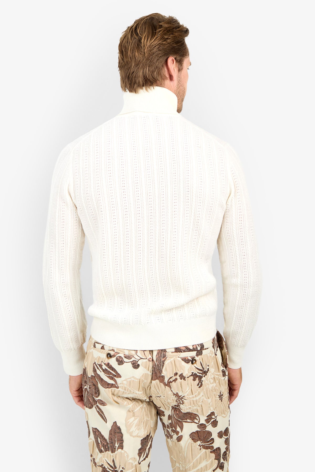 A man faces away, wearing the Oberon Turtleneck Sweater - Merino Wool in white with beige floral-patterned pants, set against a plain white background.