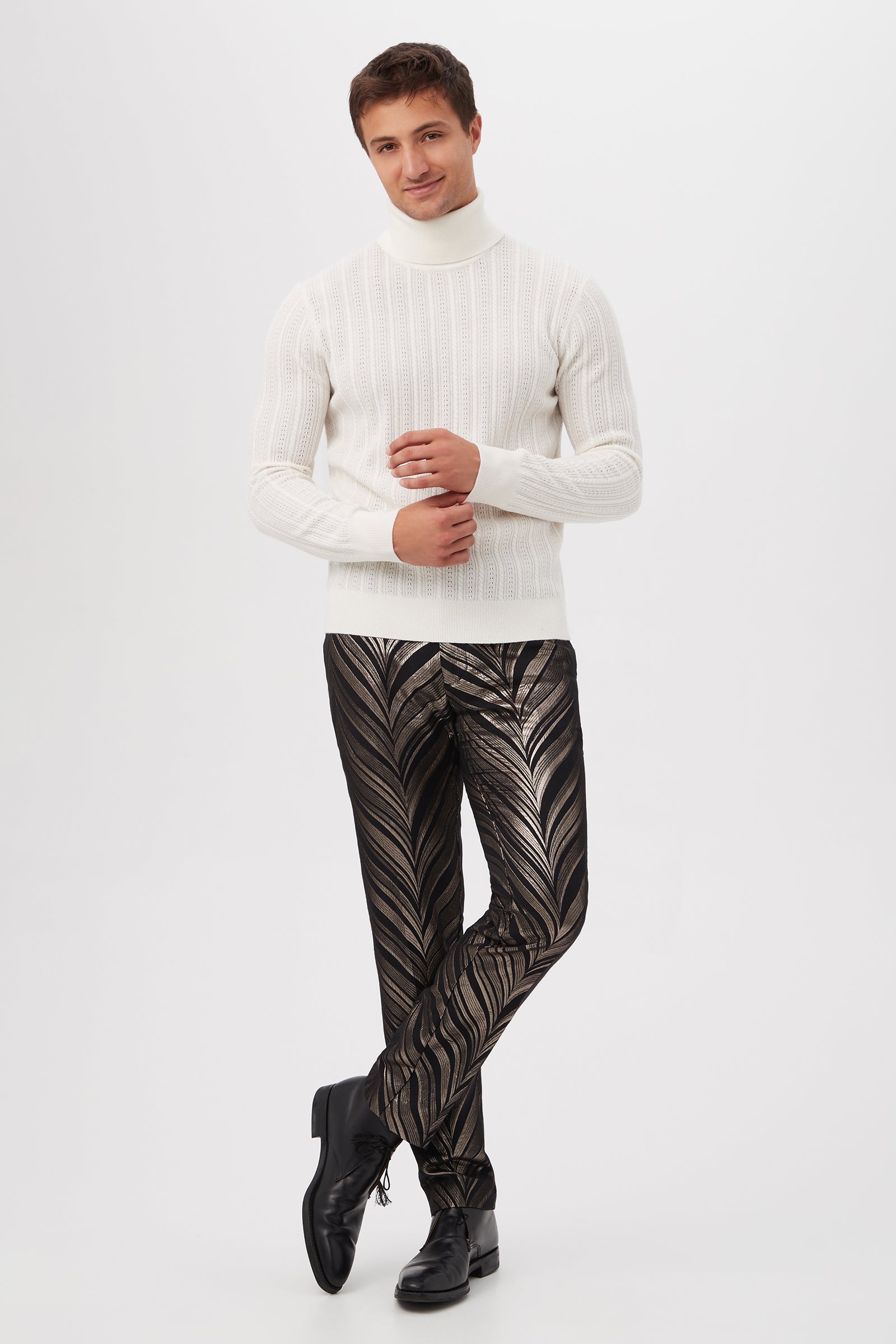 A man with short hair smiles as he poses with crossed legs and hands together, wearing the Oberon Turtleneck Sweater - Merino Wool and black pants featuring a bold, shiny leaf-like pattern against a white background.