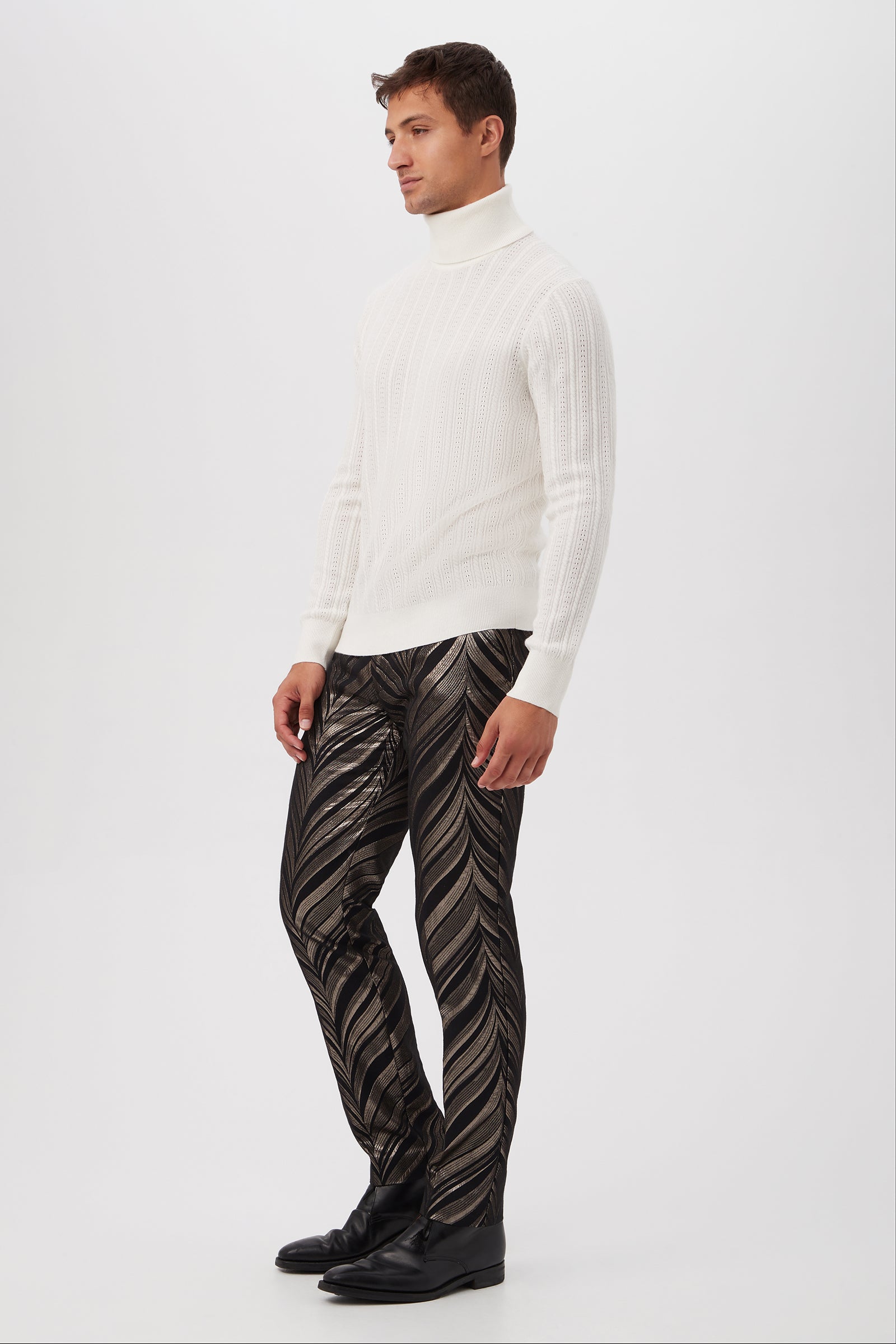 A man stands against a white background wearing the Oberon Turtleneck Sweater in merino wool, metallic black and gold patterned pants, and black shoes. He looks to the side with a neutral expression.