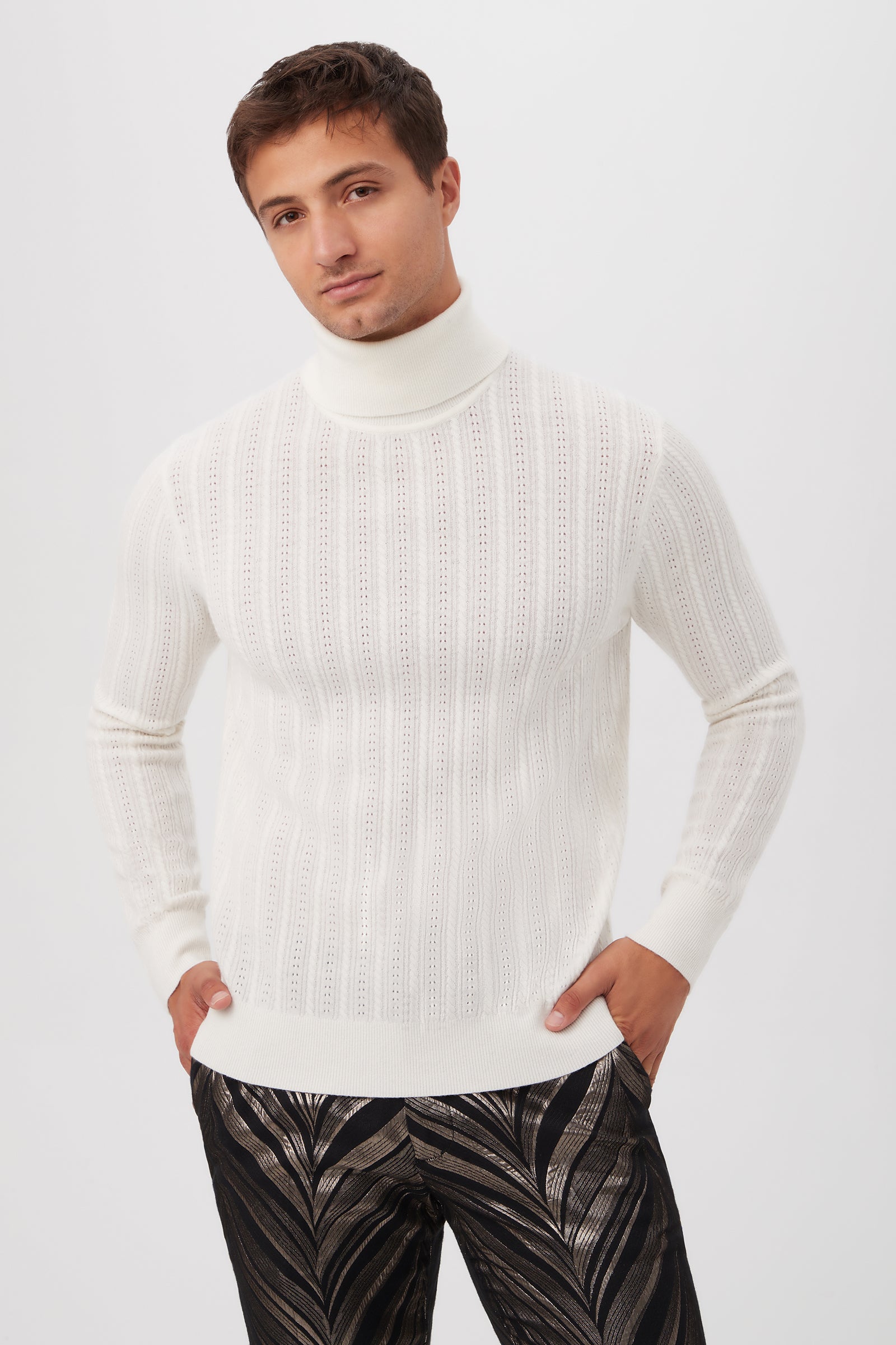 A man wears the Oberon Turtleneck Sweater in Winter White merino wool and black metallic leaf pattern pants, standing against a plain light background with his hands in his pockets.