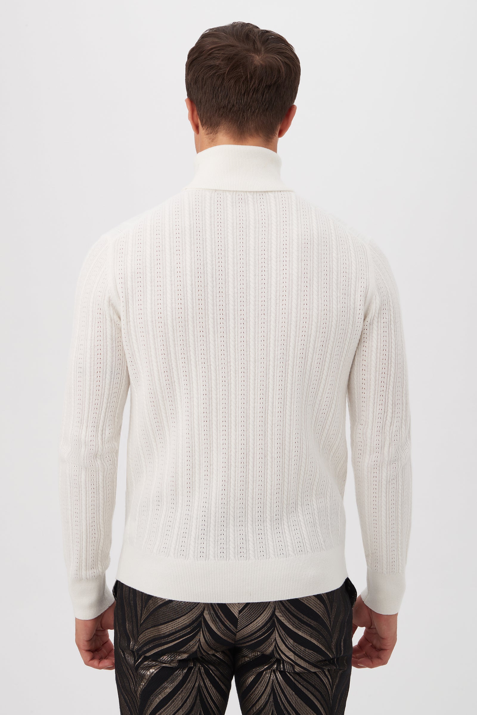 A man stands with his back to the camera, wearing the Oberon Turtleneck Sweater—crafted from merino wool—in white textured knit, paired with dark abstract-patterned pants against a plain white background.