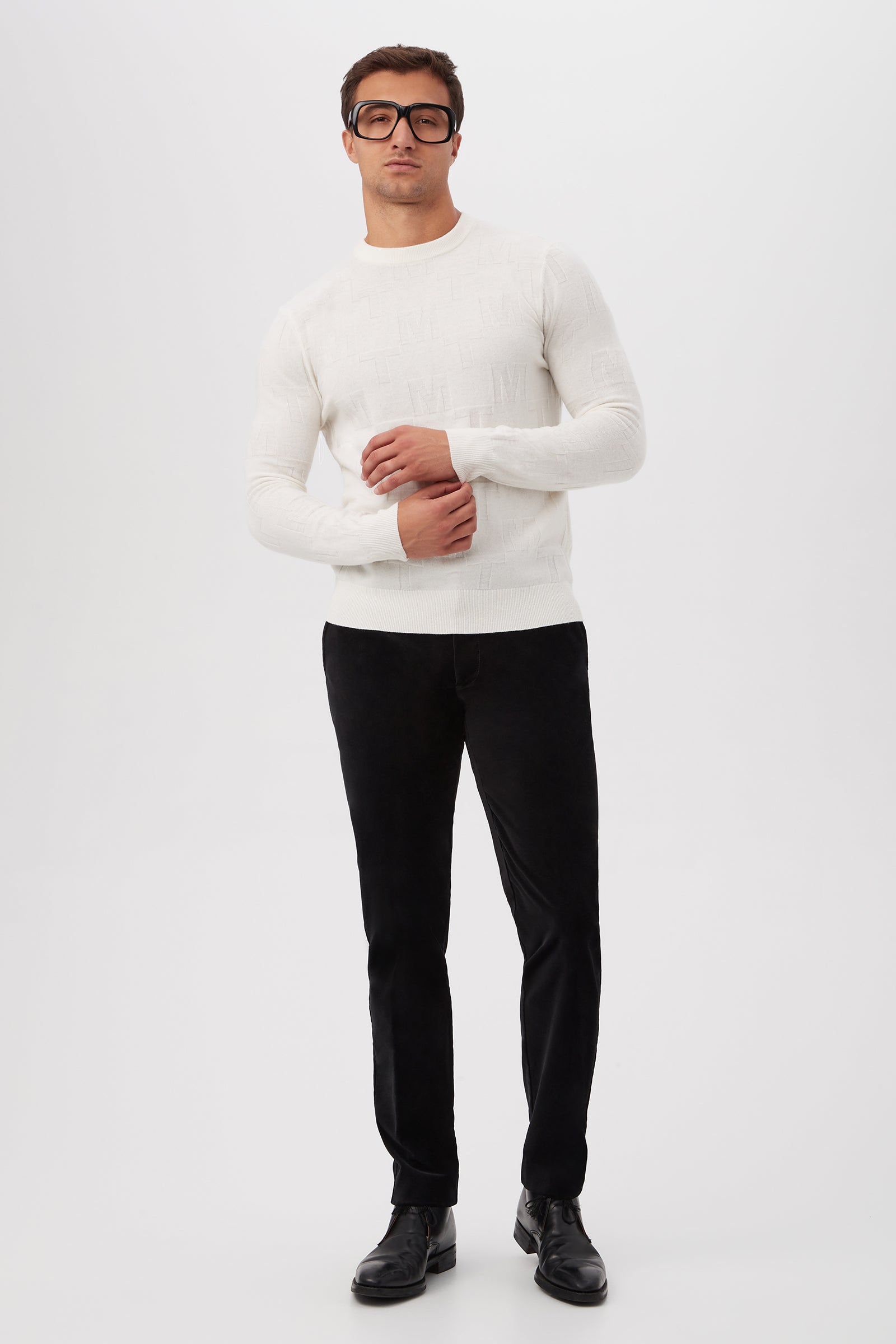A man stands against a plain white background with his hands lightly clasped, wearing black glasses and the Mr Turk Crewneck Sweater - Merino Wool in white, paired with black pants and black shoes.