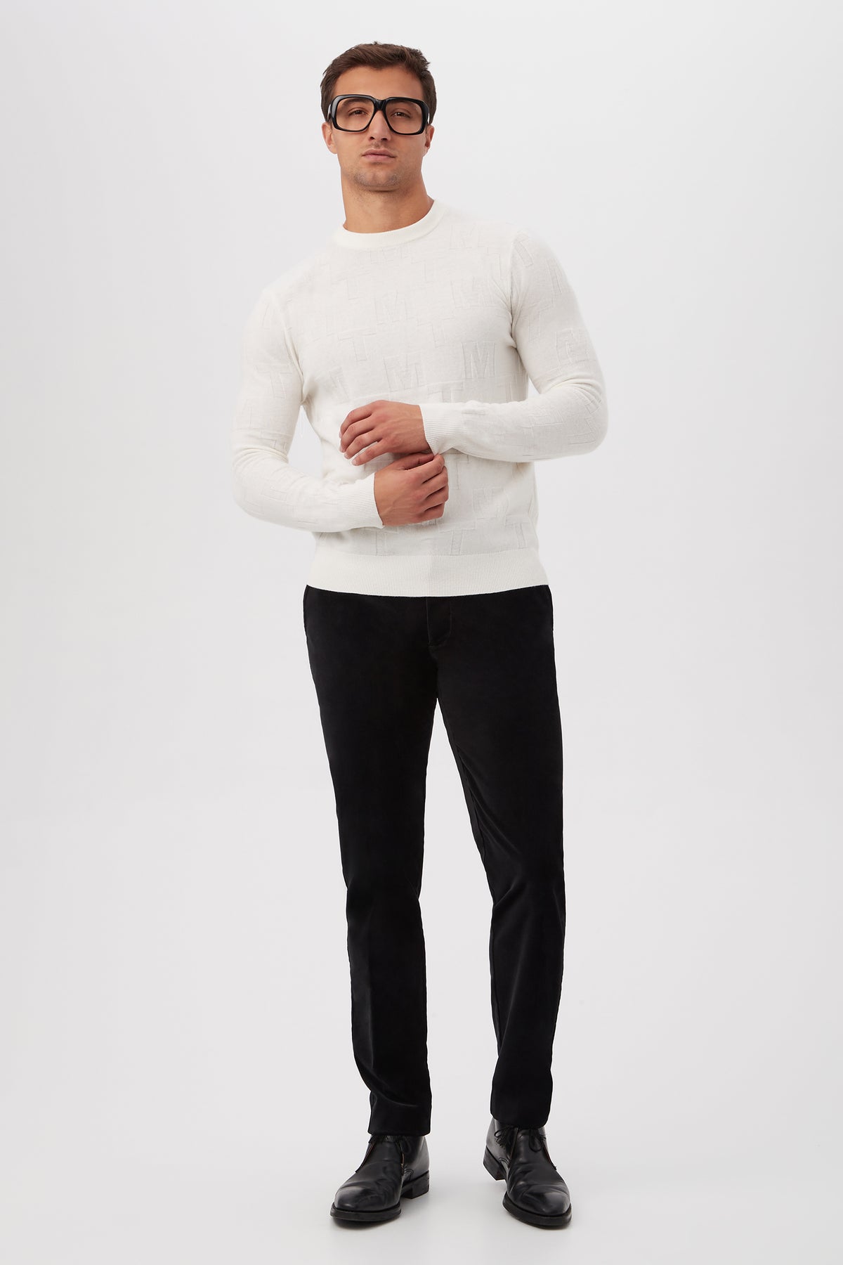 A man stands against a plain white background with his hands lightly clasped, wearing black glasses and the Mr Turk Crewneck Sweater - Merino Wool in white, paired with black pants and black shoes.