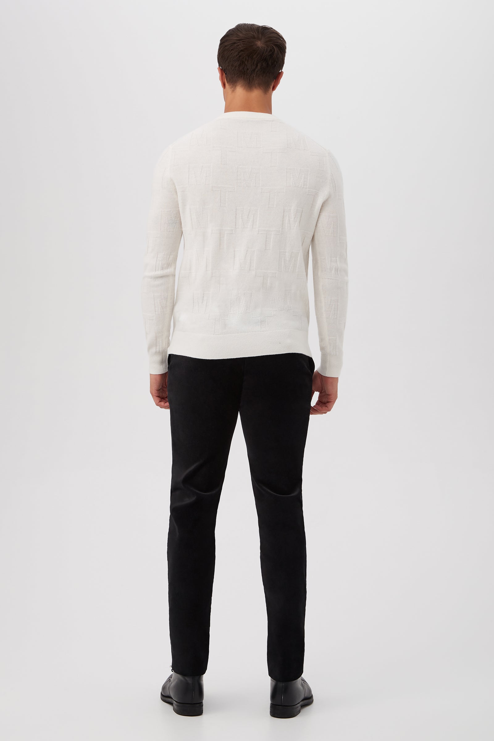 A man stands facing away from the camera, wearing the Mr Turk Crewneck Sweater in white merino wool, paired with black pants and black shoes against a plain white background.