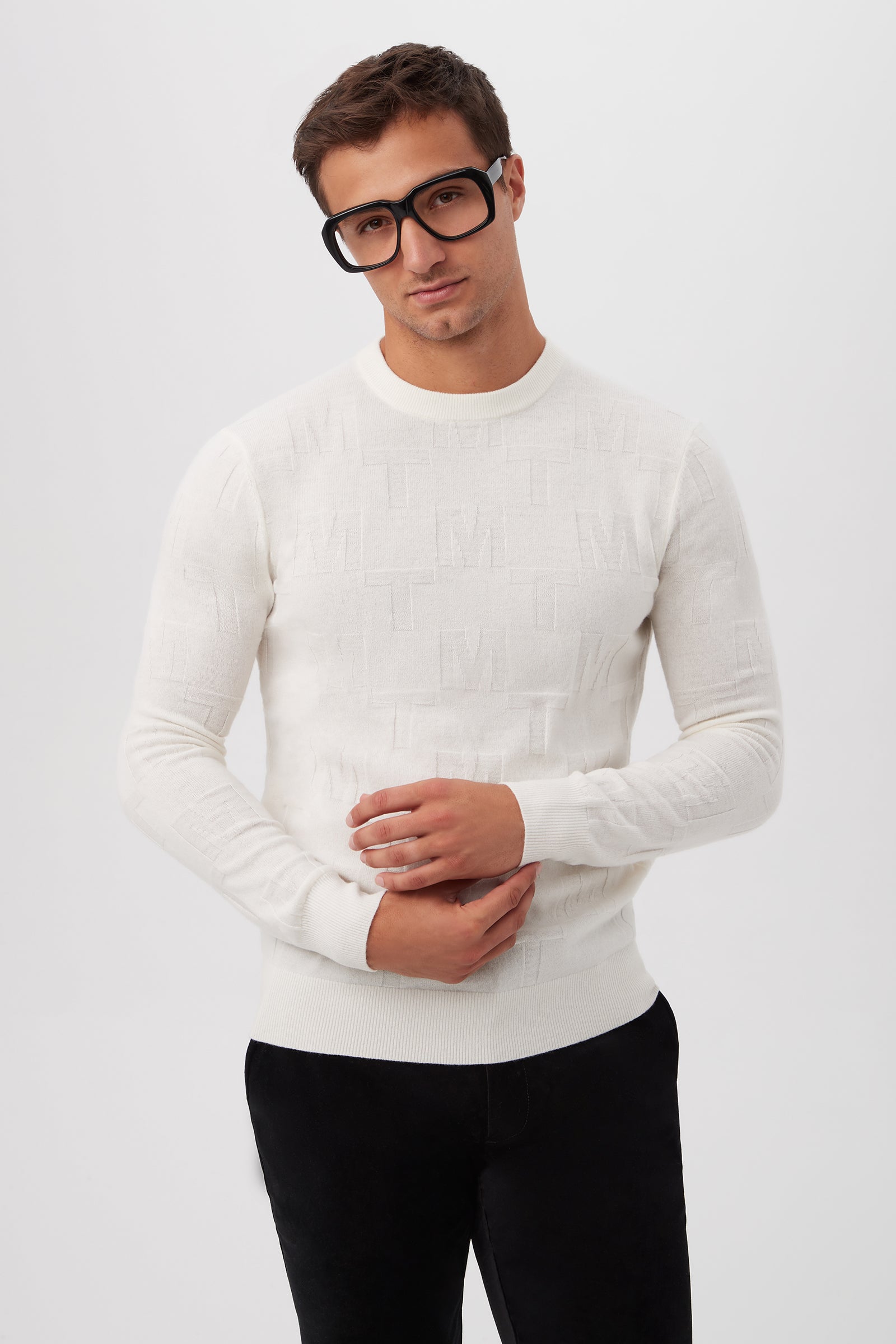 A man with short brown hair and black-rimmed glasses wears the Mr Turk Crewneck Sweater in Winter White merino wool with a subtle embossed pattern and dark pants, standing against a plain white background.