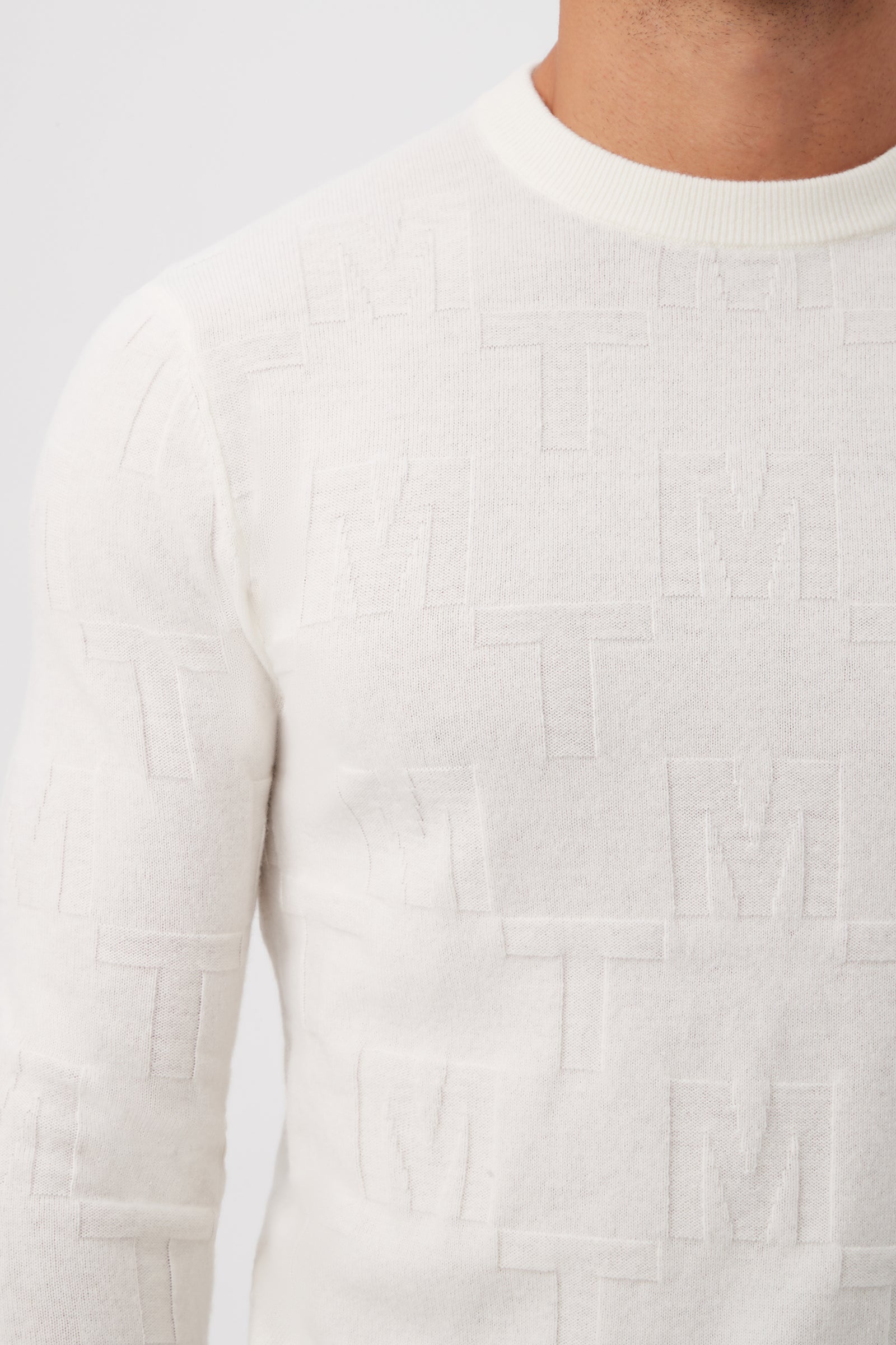 A close-up of a person wearing the Mr Turk Crewneck Sweater - Merino Wool, featuring textured white fabric with raised M and T letters. The plain background and cropped frame keep the face out of view.