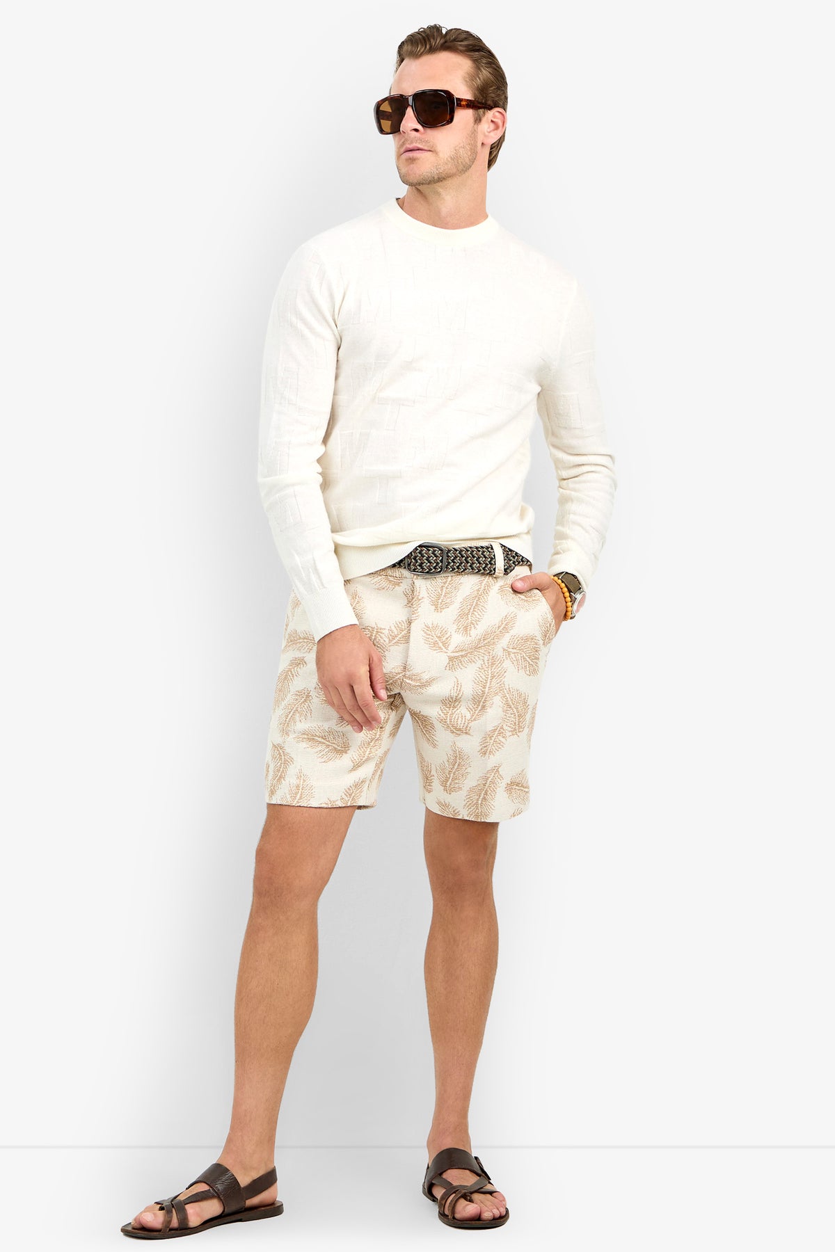 A man wearing sunglasses, the Mr Turk Crewneck Sweater - Merino Wool, patterned shorts, a belt, and sandals stands relaxed against a plain background.