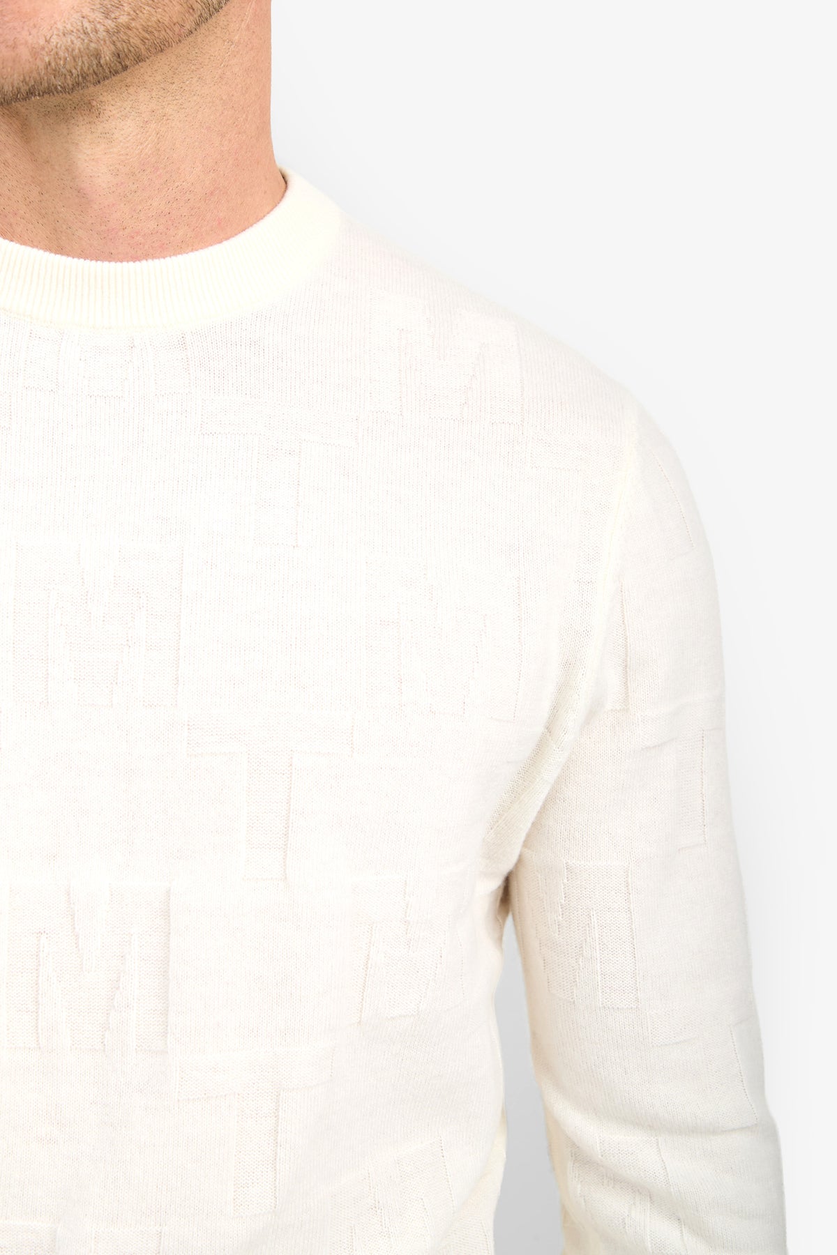 A close-up of a person wearing the Mr Turk Crewneck Sweater - Merino Wool, showcasing its textured off-white fabric and subtle embossed letter pattern. Only the lower face, neck, and upper body are visible.