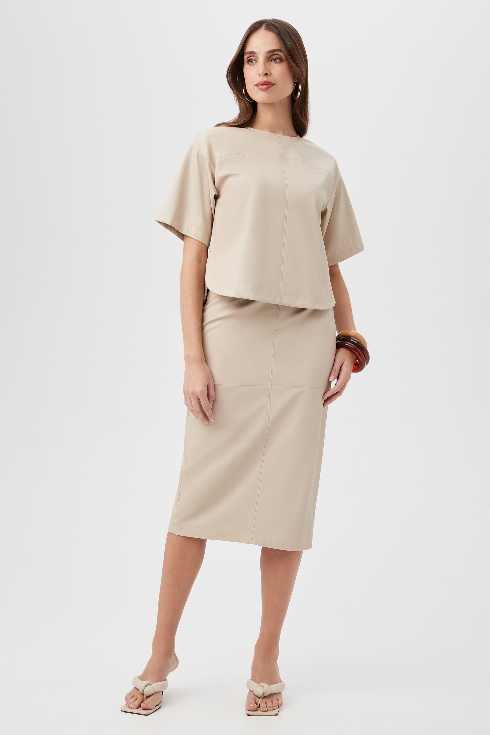 A woman with long brown hair stands against a white background, wearing the Serena Skirt—a beige, knee-length skirt—paired with a matching short-sleeve top, heeled sandals, hoop earrings, and bangle bracelets.