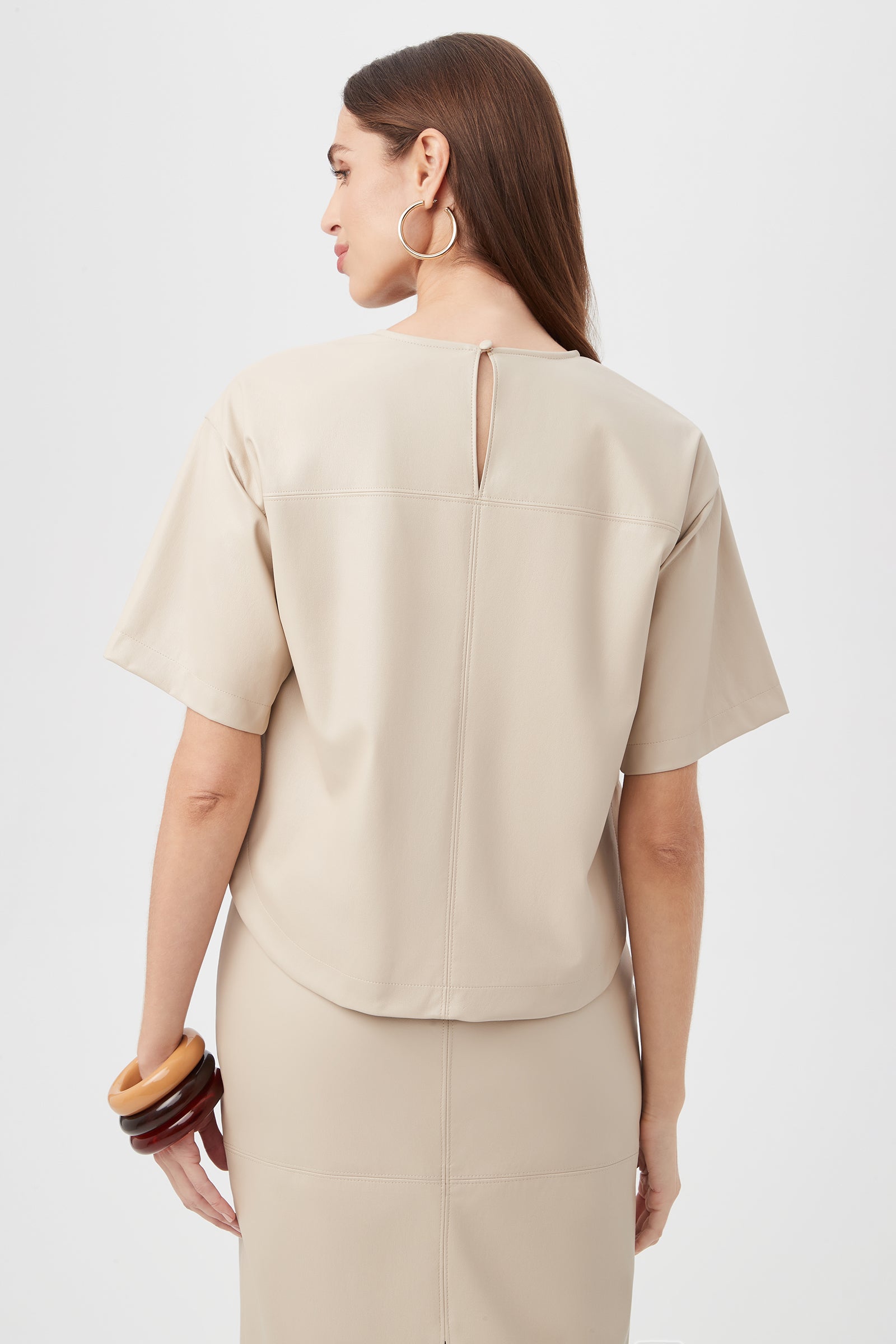 Woman with straight brown hair seen from the back, wearing the Moira Top in beige with short sleeves and a small keyhole at the neck, paired with a matching skirt. She accessorizes with large hoop earrings and chunky bracelets.