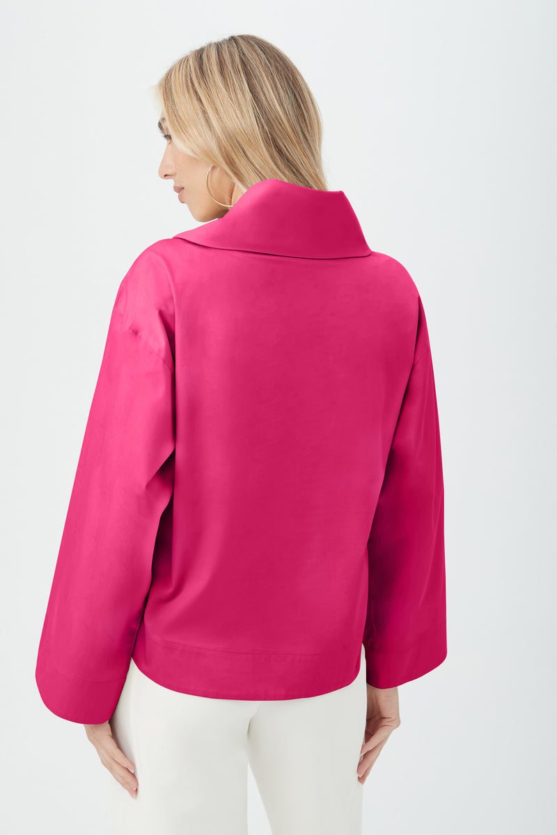 A woman with blonde hair is seen from the back, wearing the Tomie Top—a bright pink, long-sleeve jacket with a wide collar and loose fit—paired with white pants against a plain light background.