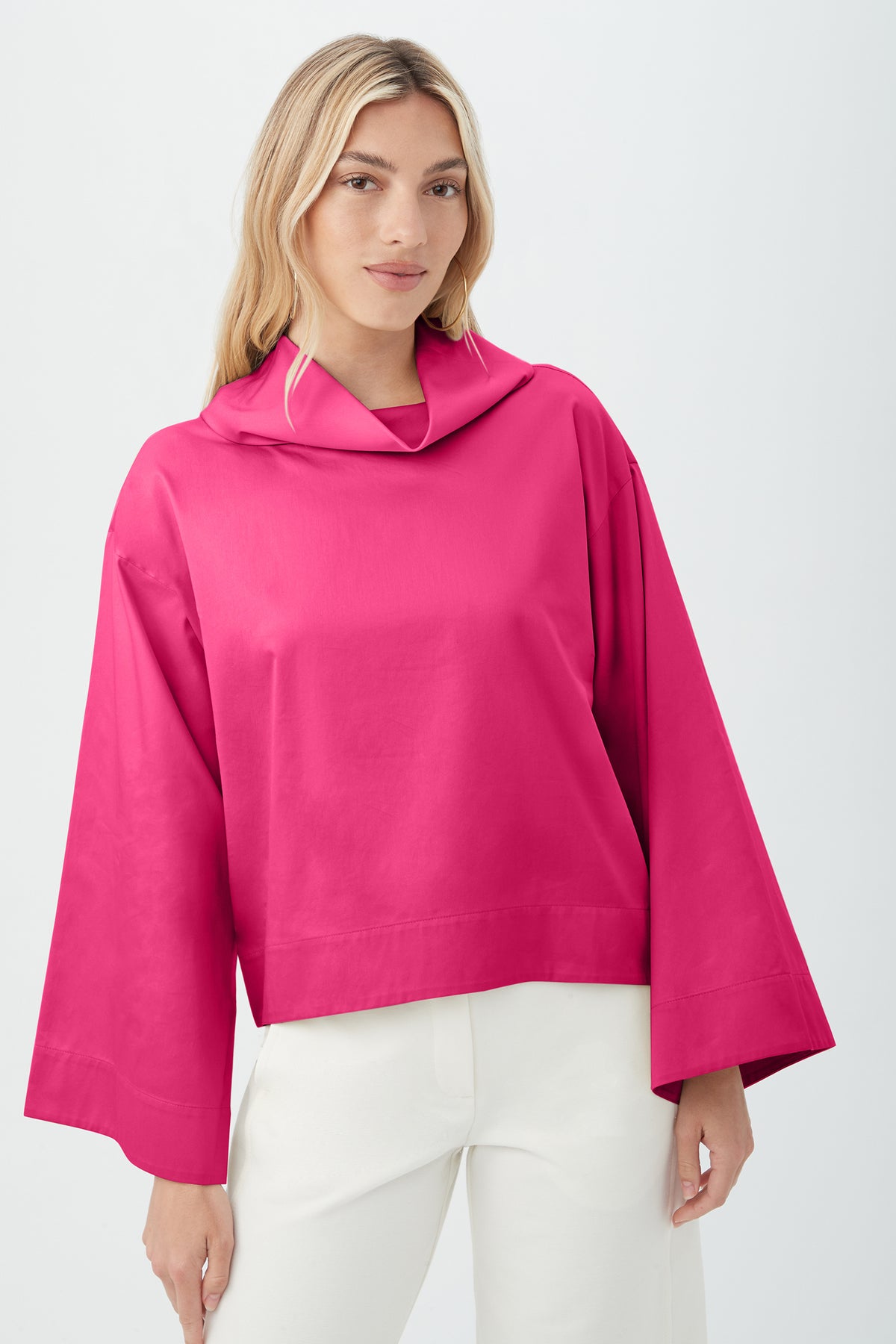 A woman with long blonde hair wears the Tomie Top in Petal Pink, a loose-fitting, long-sleeve top with wide sleeves and a cowl neckline, paired with white pants against a plain light background.