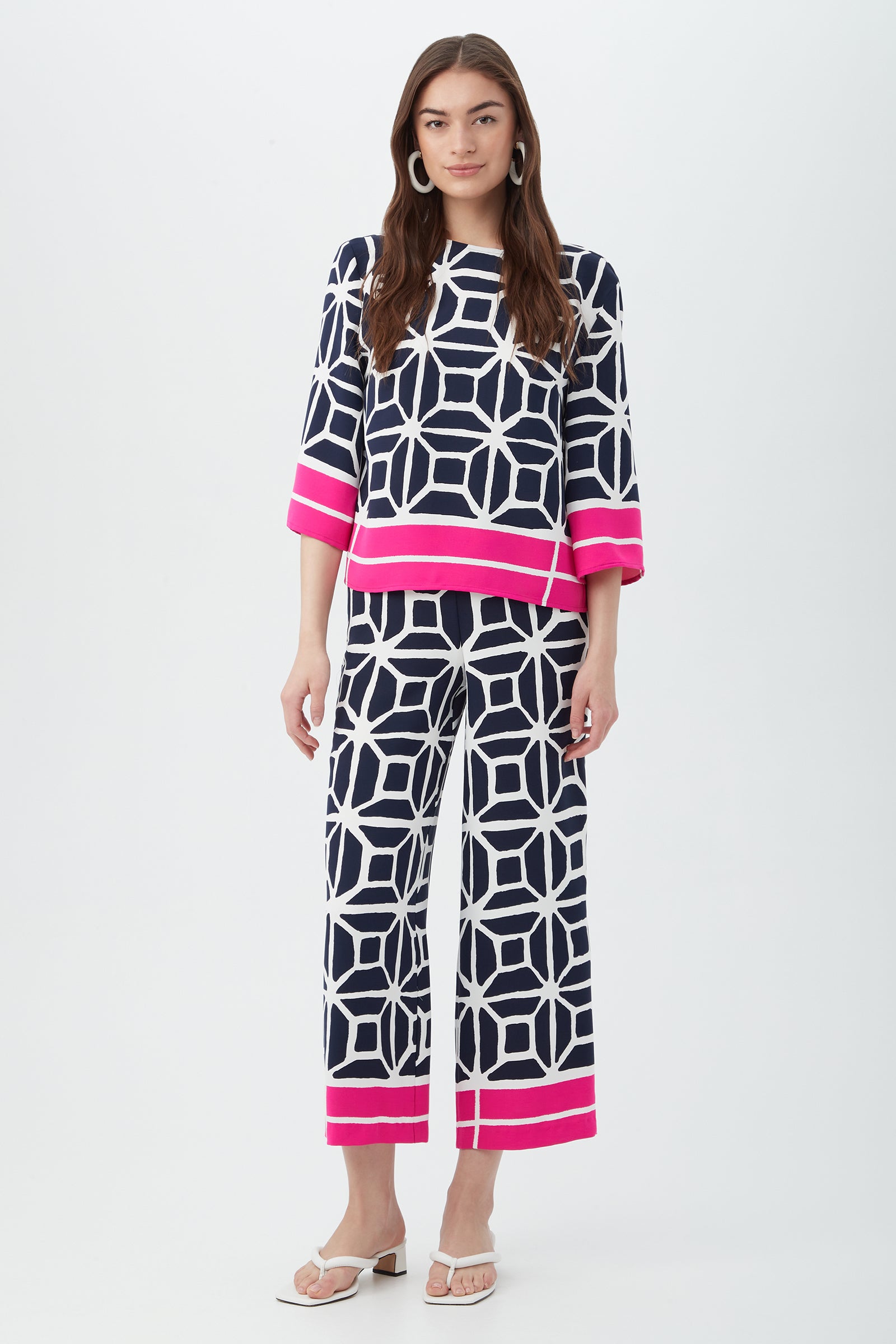 A woman wears the Orlett Wide-Leg Crop Pant in Durell Georgette, paired with a matching navy and white geometric print top featuring bright pink borders. She has loose brown hair, white earrings, and white sandals.